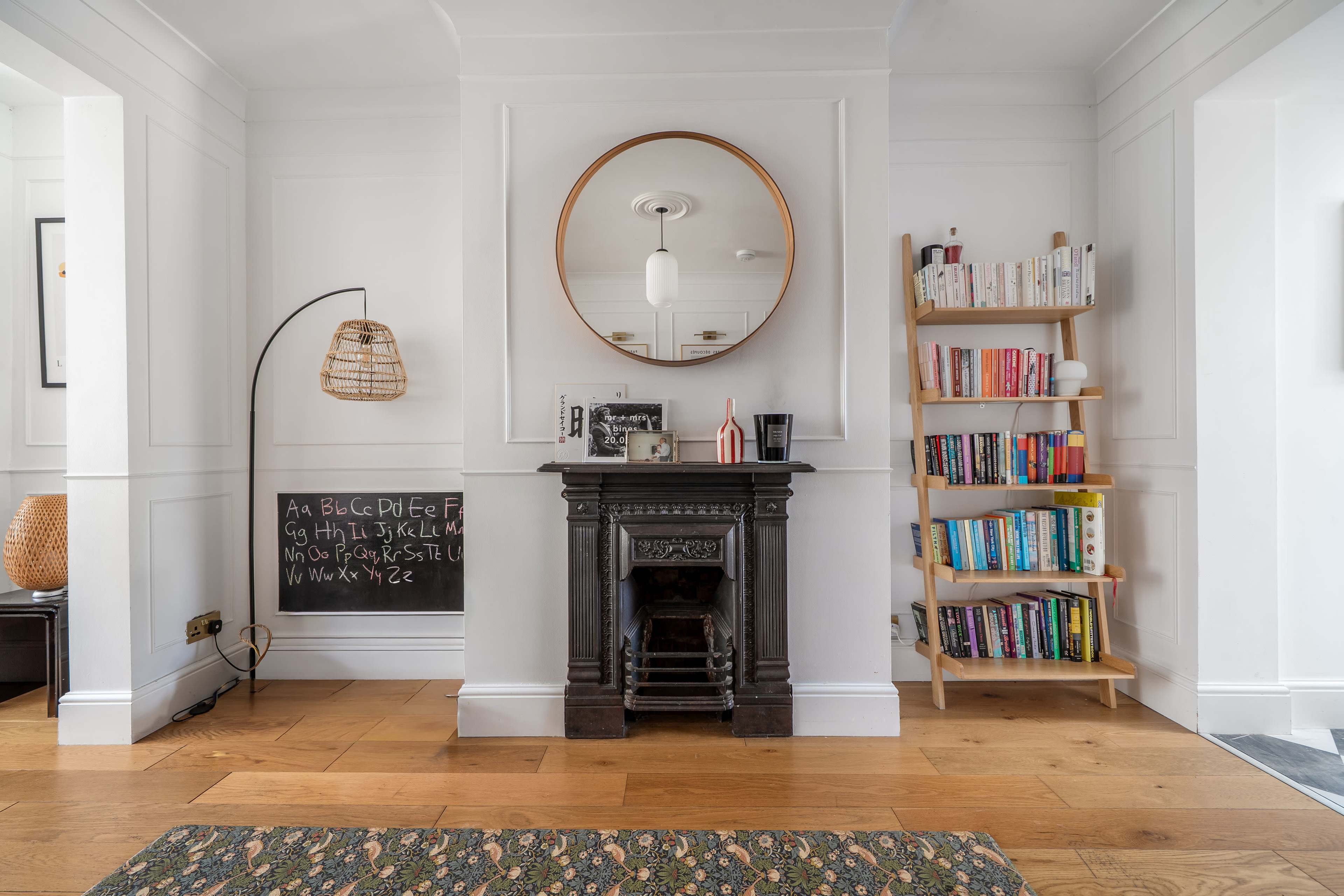 The image shows a living room featuring a vintage fireplace, a round mirror above it, a bookshelf filled with books, a wall-mounted chalkboard, and wooden flooring.