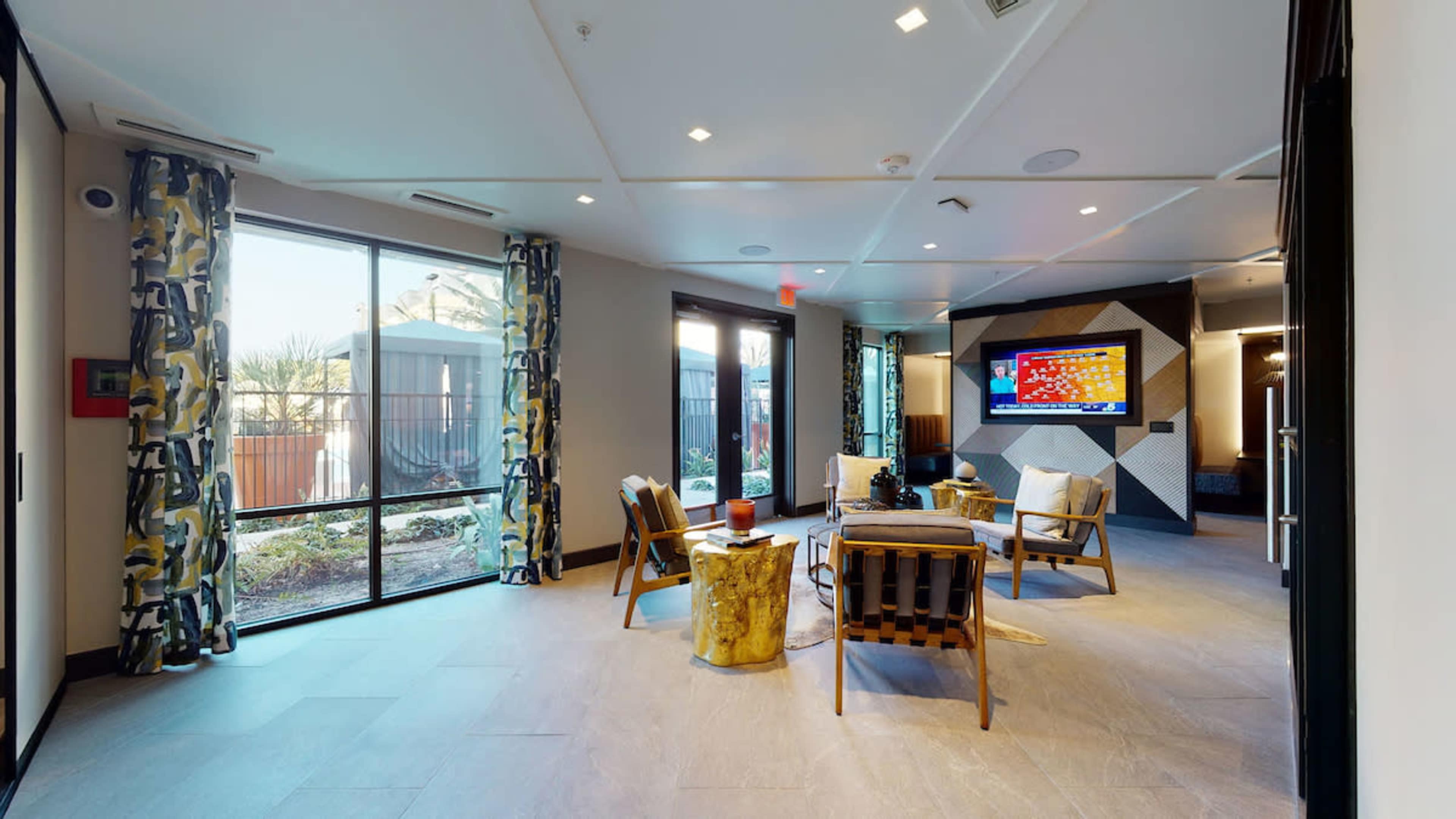 A modern lounge area with large windows, featuring seating arrangements, a wooden table, and a wall-mounted television.