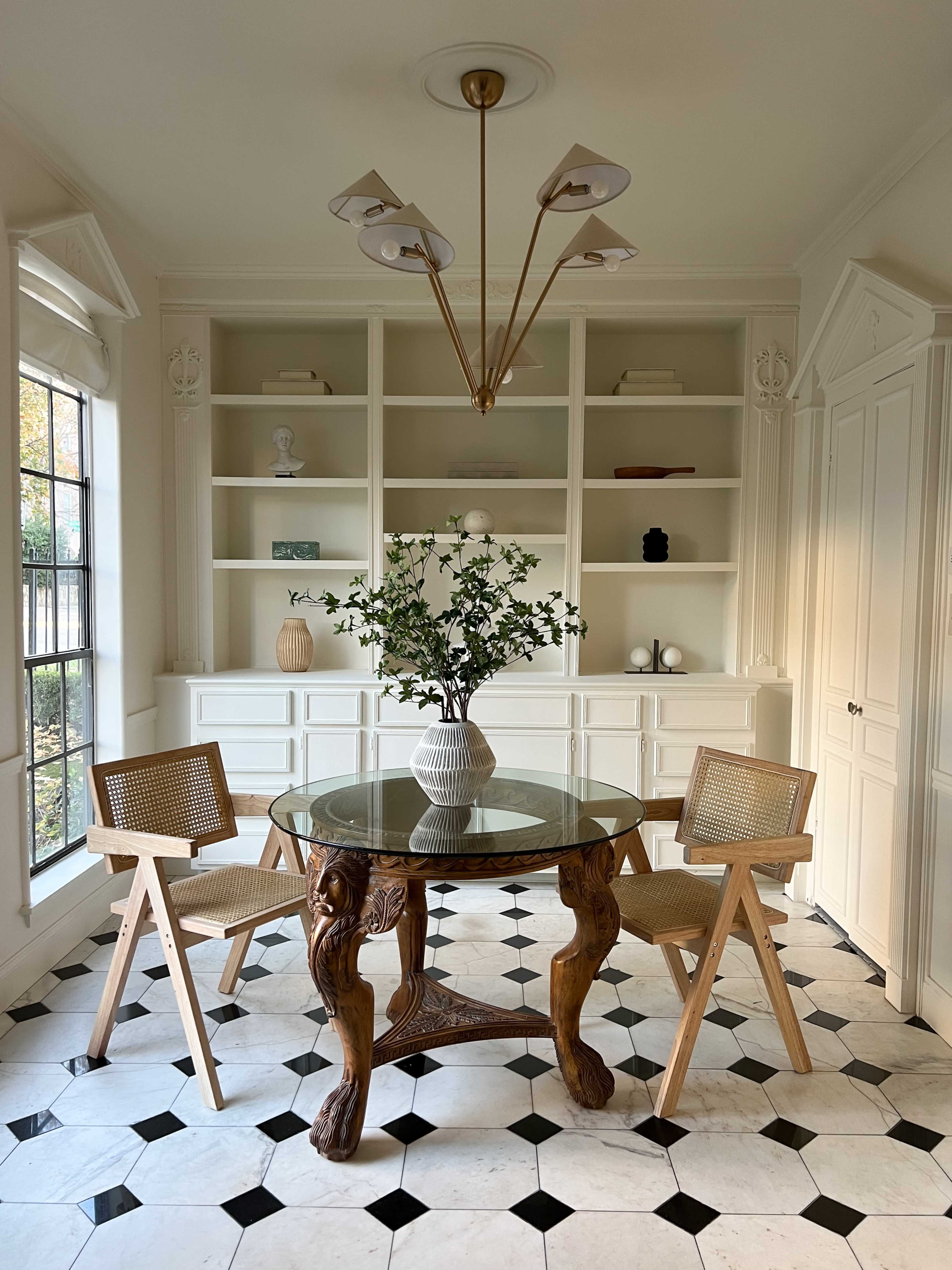 A dining area features a round glass table with a decorative base, surrounded by two wooden chairs, and a vase of greenery on the table, against a backdrop of white shelves and a patterned tile floor.