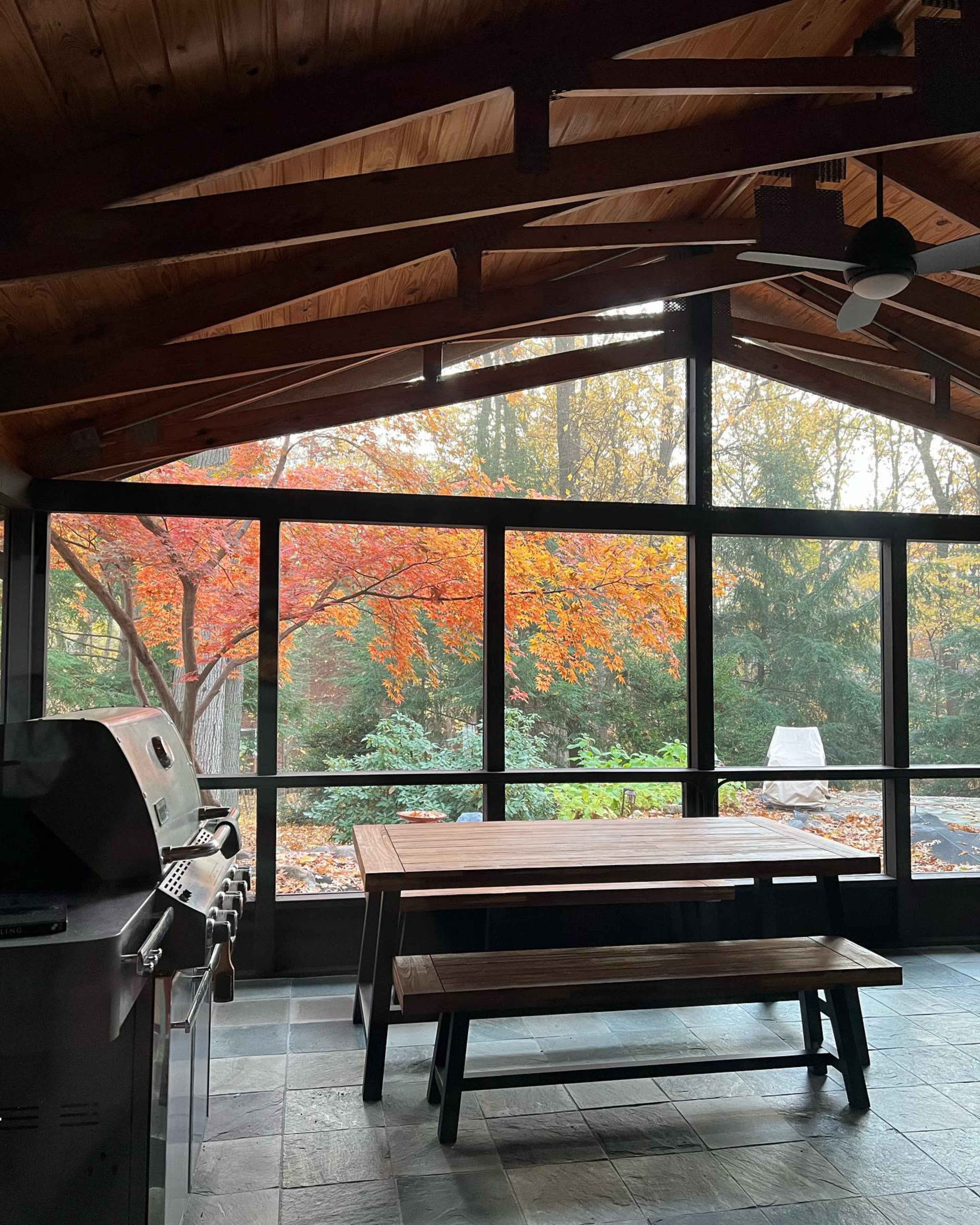 The image shows a screened-in area with a wooden table and an outdoor grill, overlooking a vibrant autumn landscape with colorful trees.