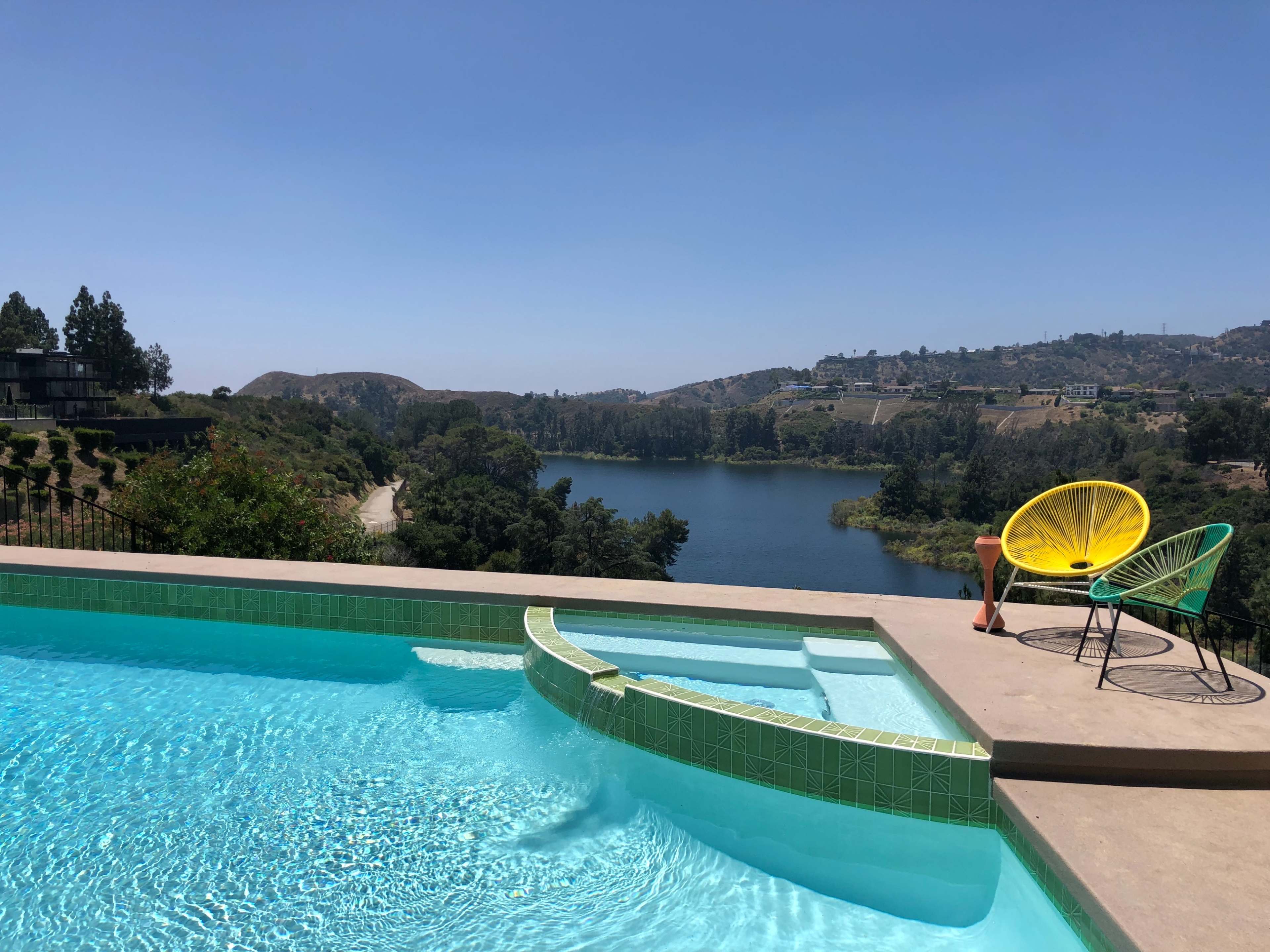 A swimming pool with turquoise water overlooks a body of water surrounded by hills and greenery.