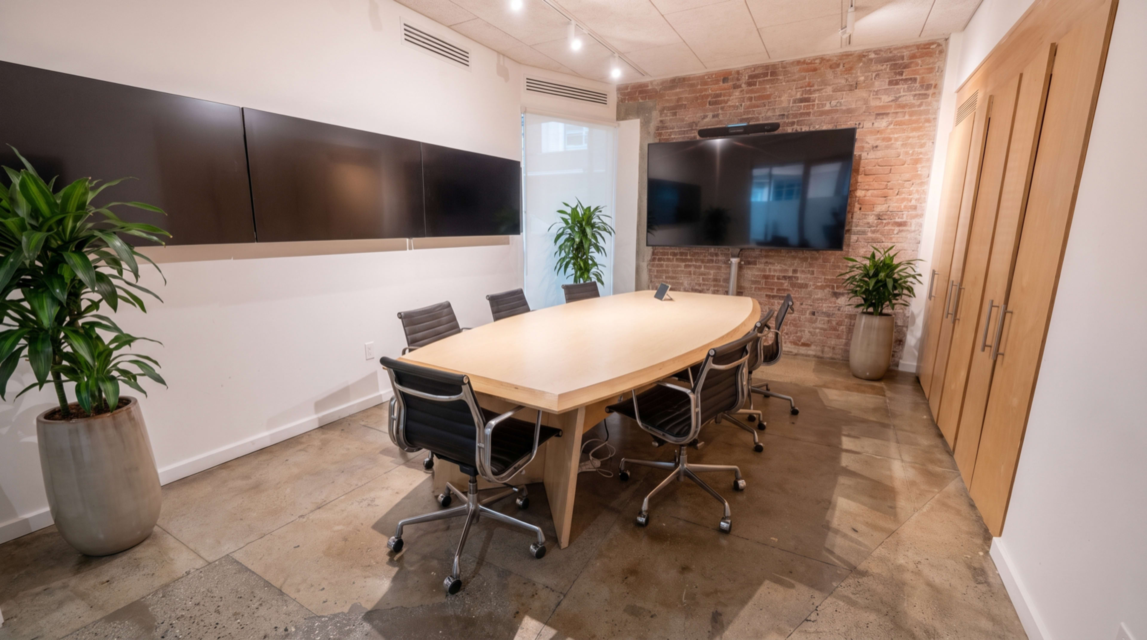 A conference room features an oval wooden table surrounded by black chairs and two large screens mounted on the walls, with brick accents and potted plants adding greenery.