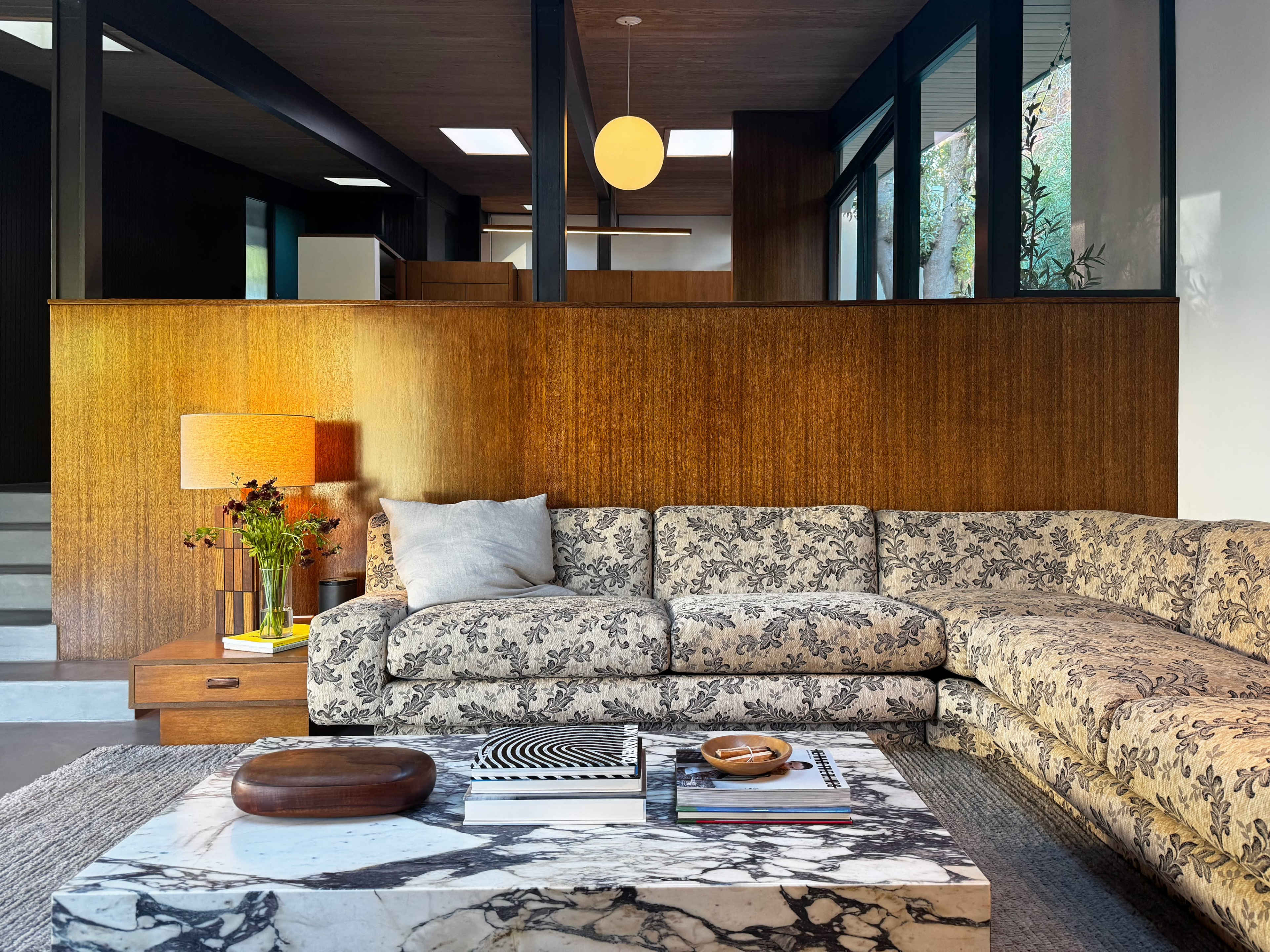 A modern living room features a floral-patterned sectional sofa, a marble coffee table, and warm wooden accents with large windows bringing in natural light.