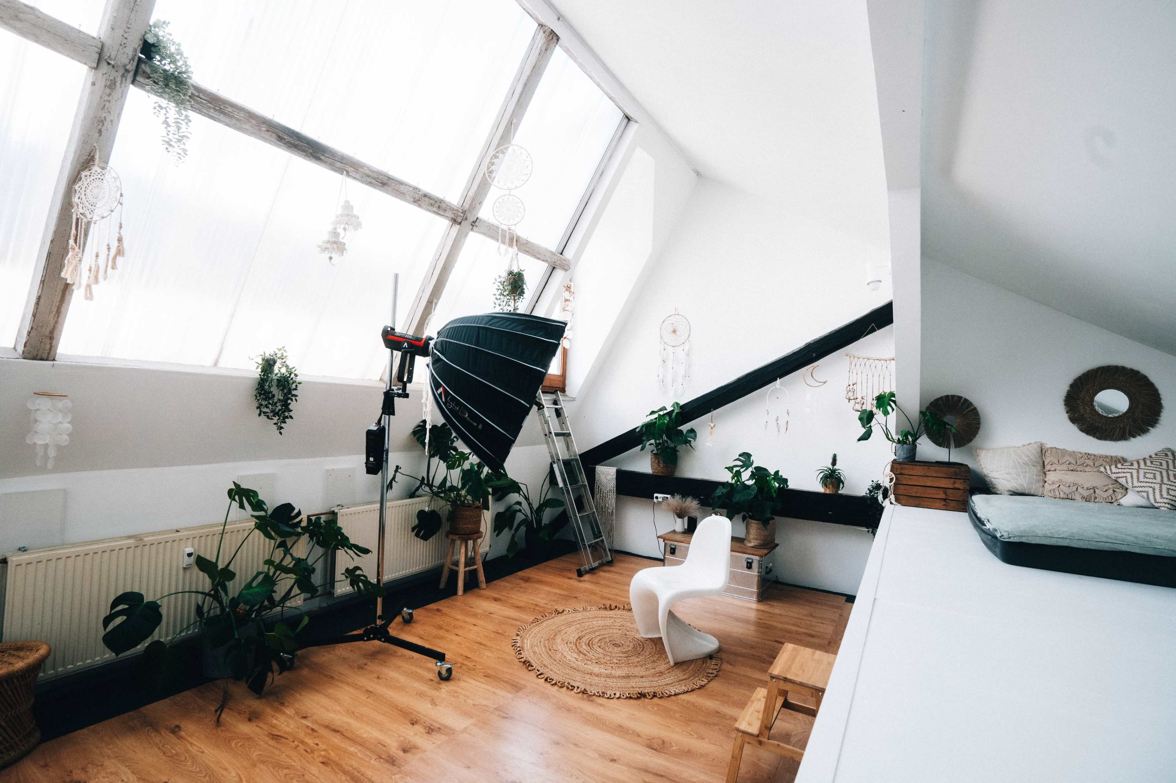 The image shows a bright, modern studio space with large windows, a photography light setup, plants, and minimalist furniture.