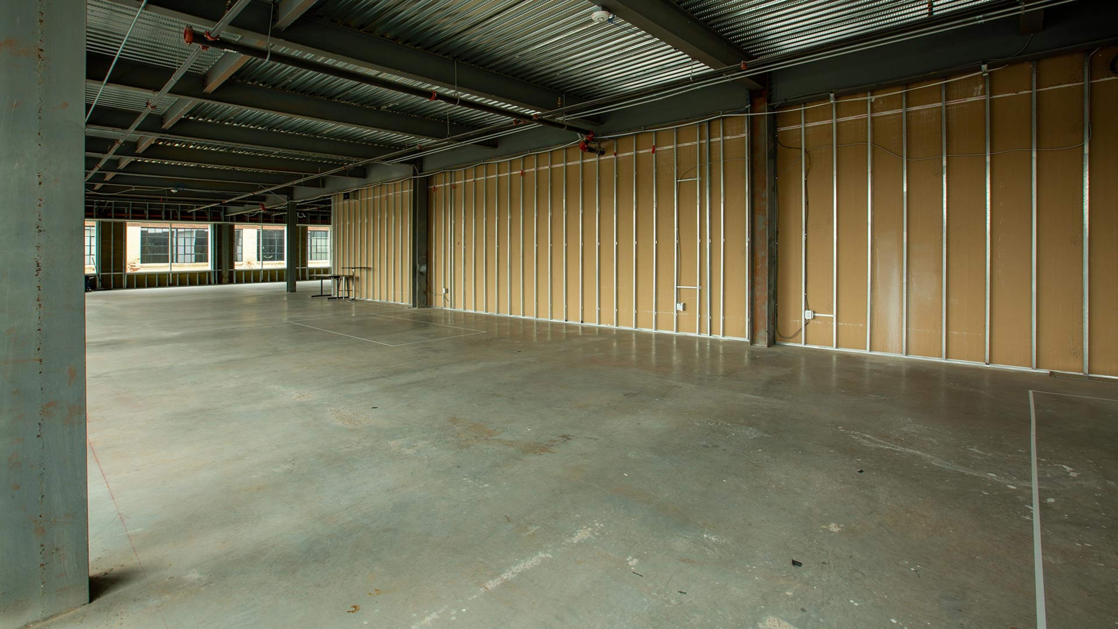 An empty, industrial-looking interior space with concrete floors and exposed structural beams, featuring bare walls with metal framing.