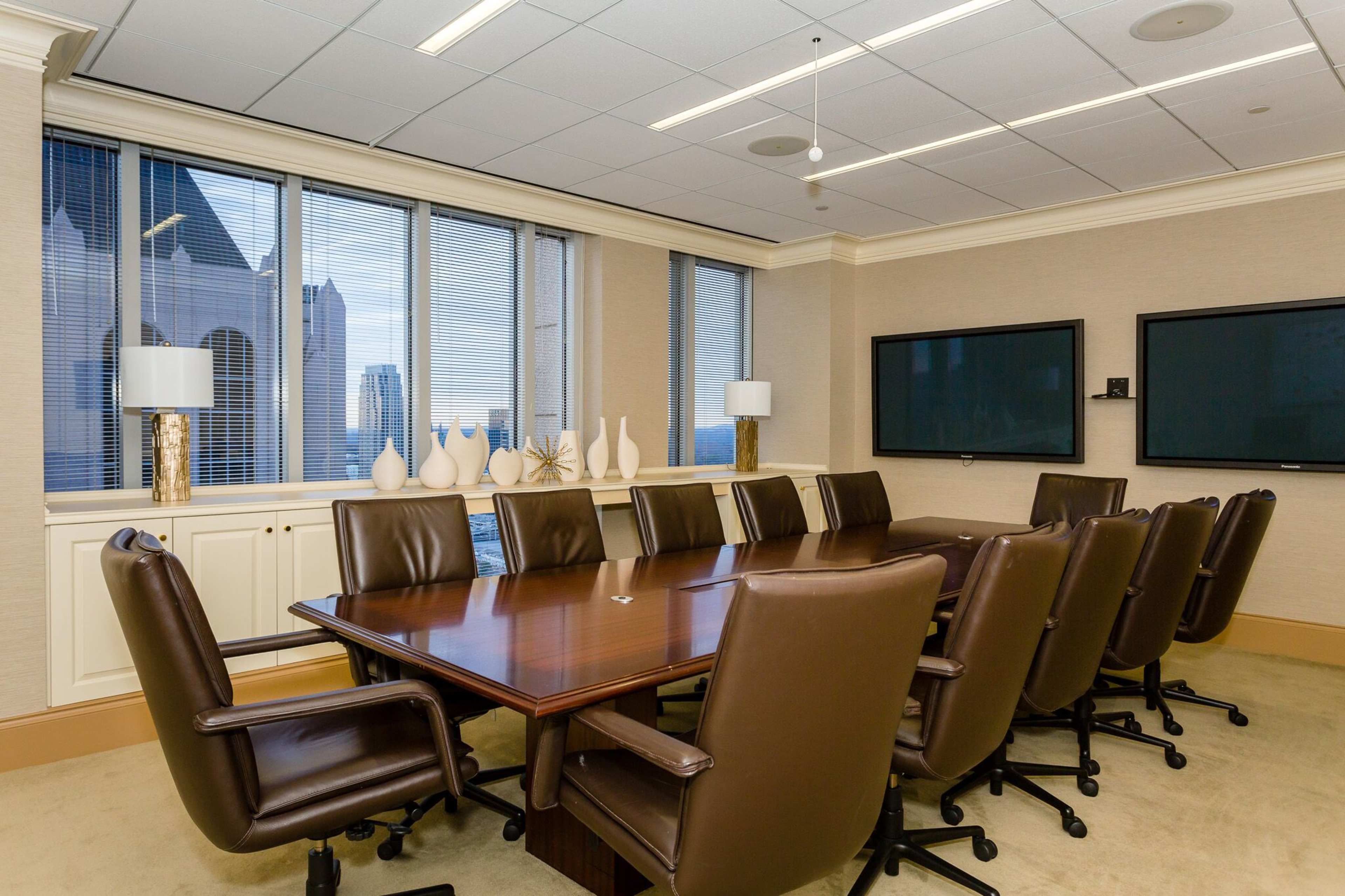 A spacious conference room features a large wooden table surrounded by leather chairs, with windows overlooking a cityscape and two wall-mounted screens.