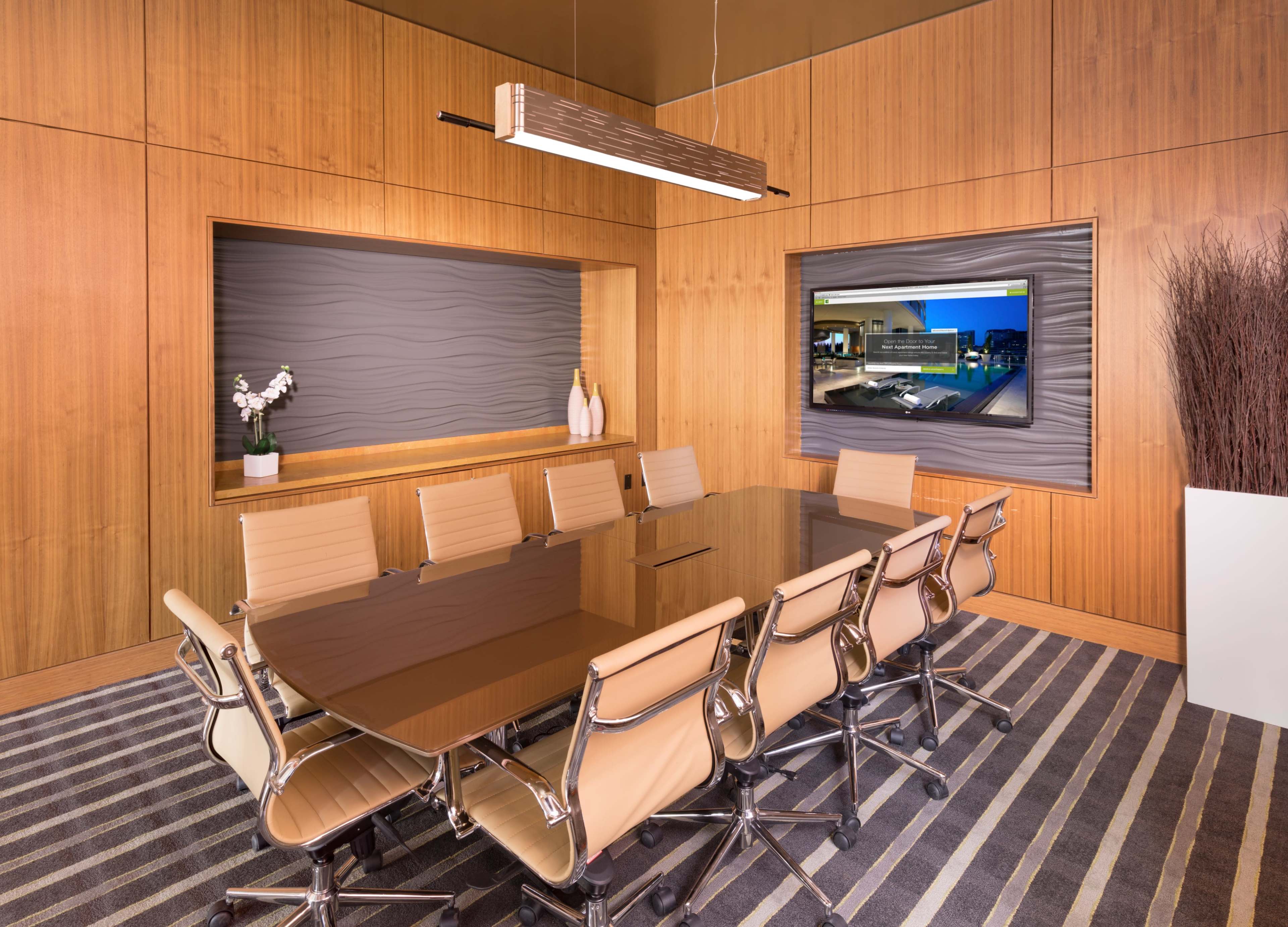 The image shows a modern conference room with a large wooden table surrounded by beige chairs, wooden paneling on the walls, and a TV screen displaying a city view.