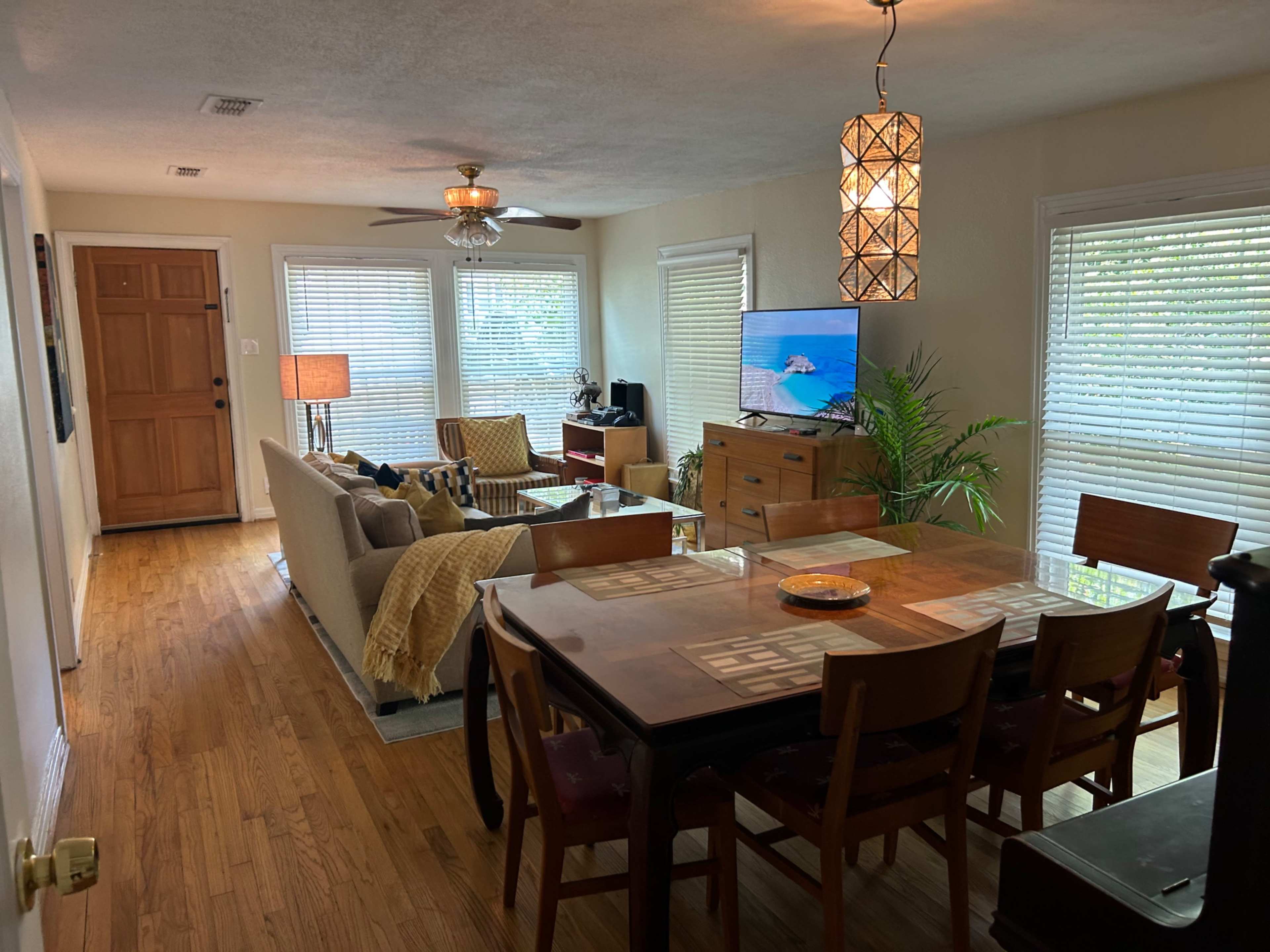 The image shows a bright living and dining area with a sofa, dining table, television, and several windows allowing natural light to enter.