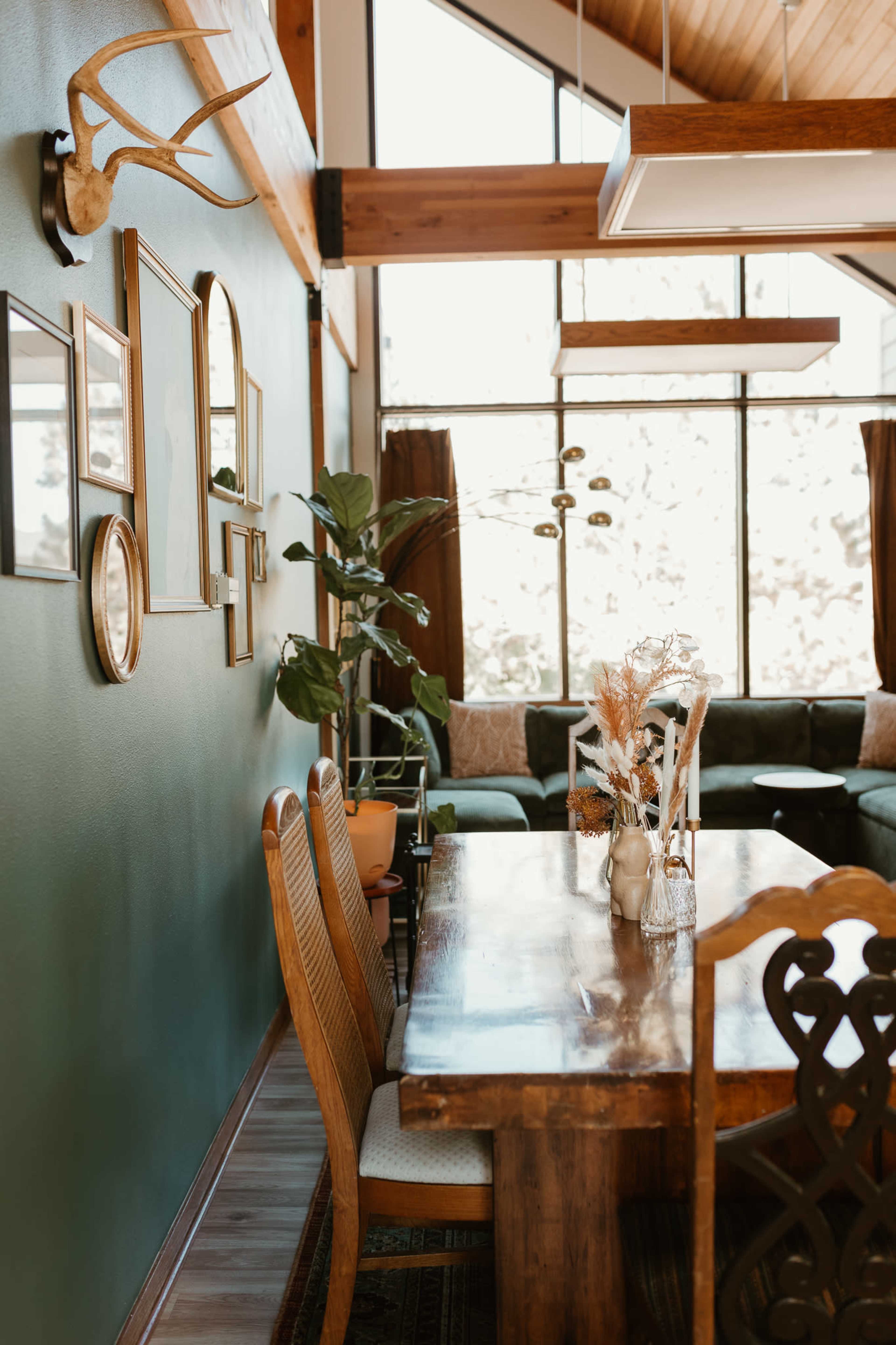 The image shows a dining area with a wooden table and chairs, side by side with a green wall featuring framed pictures and a decorative plant, leading to a spacious living area with large windows.