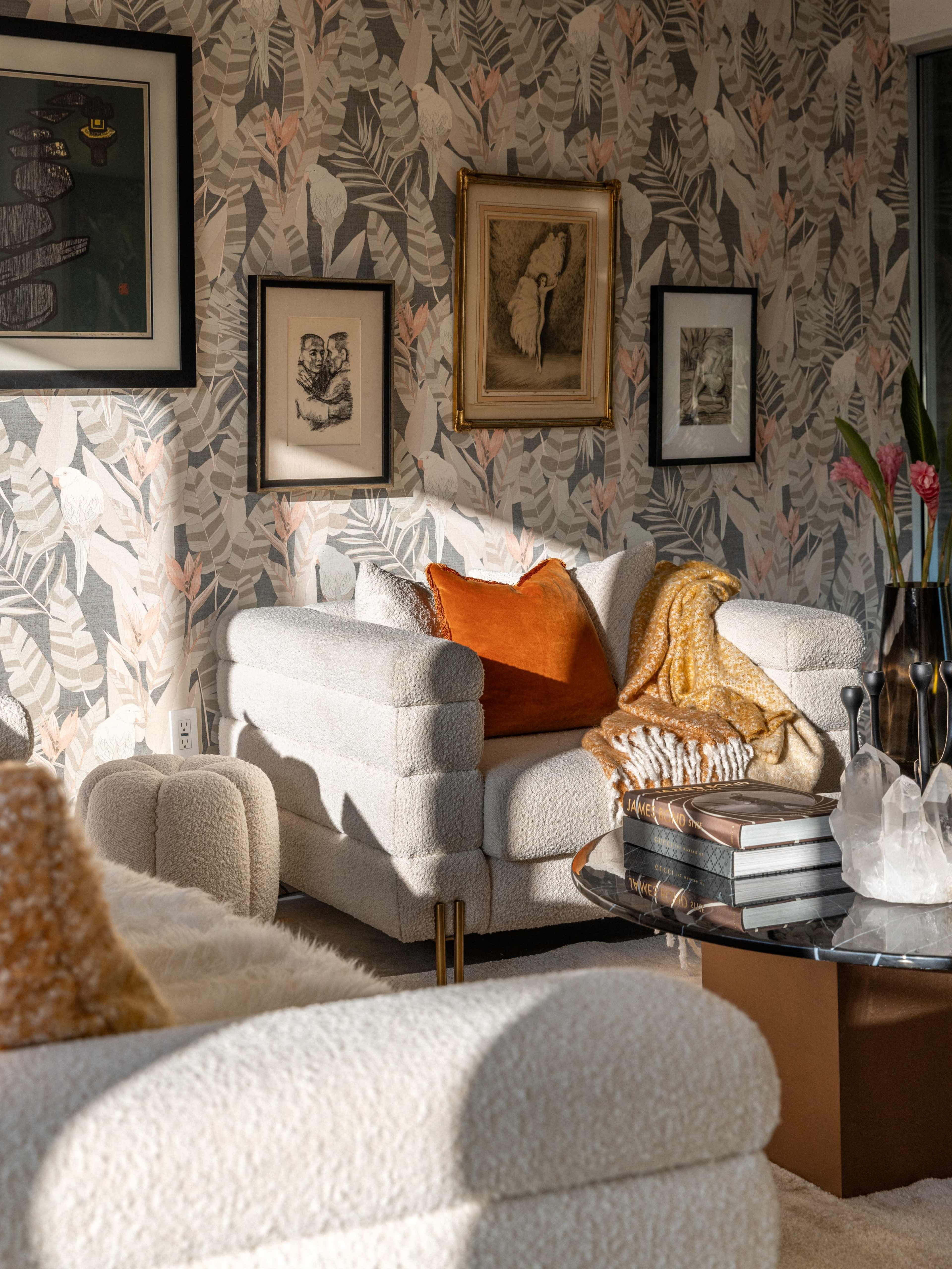 A cozy living room features a textured white sofa, an orange throw pillow, a glass coffee table with stacked books, and framed artwork on a leaf-patterned wall.