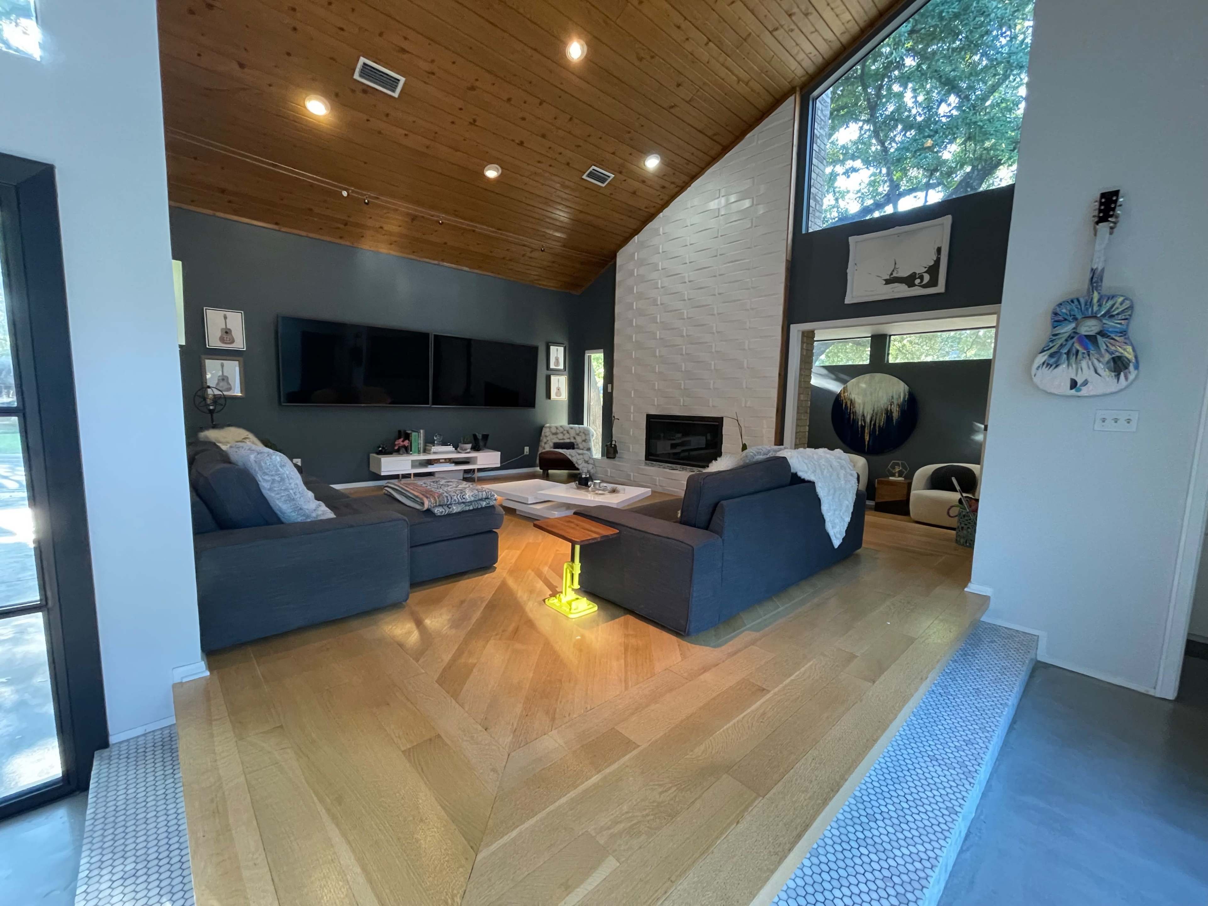 The living room features a high ceiling with wooden beams, two gray sofas arranged around a coffee table, and large windows providing natural light.