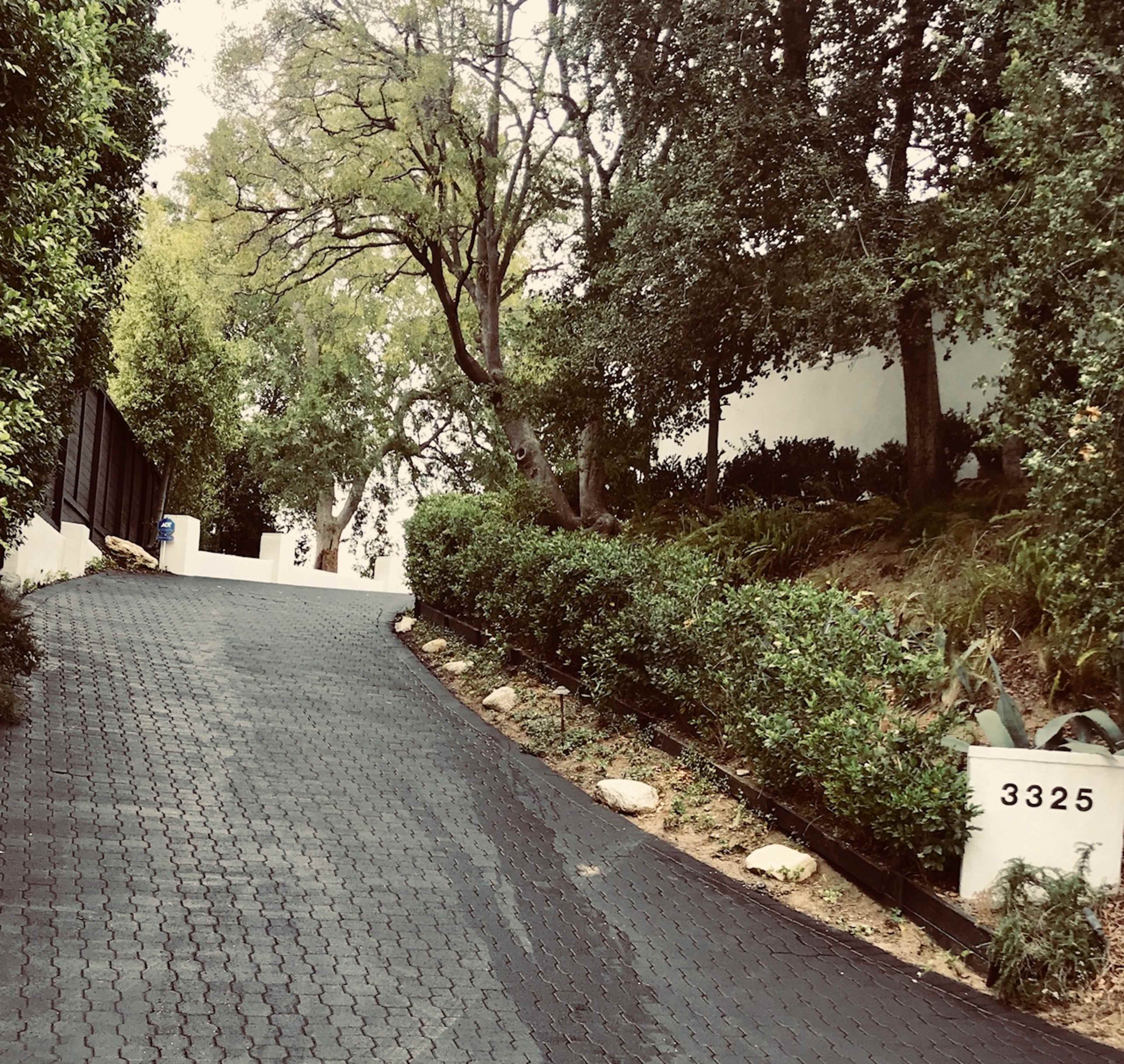 A winding driveway ascends through a landscaped area lined with trees and shrubs, leading to a property marked by the number 3325.