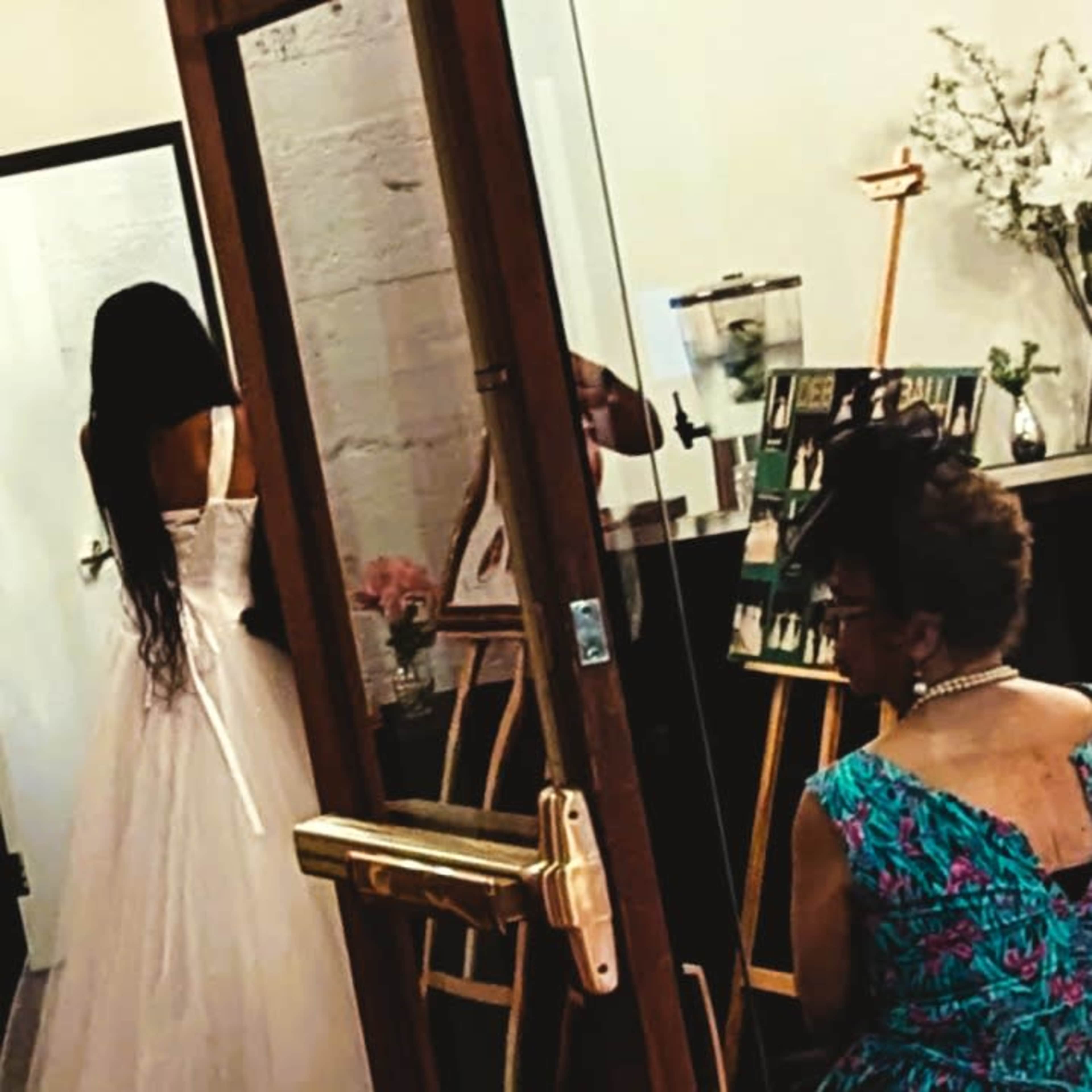A woman in a white dress stands with her back turned in front of a mirror, while another woman sits nearby, partially visible, in a patterned outfit.