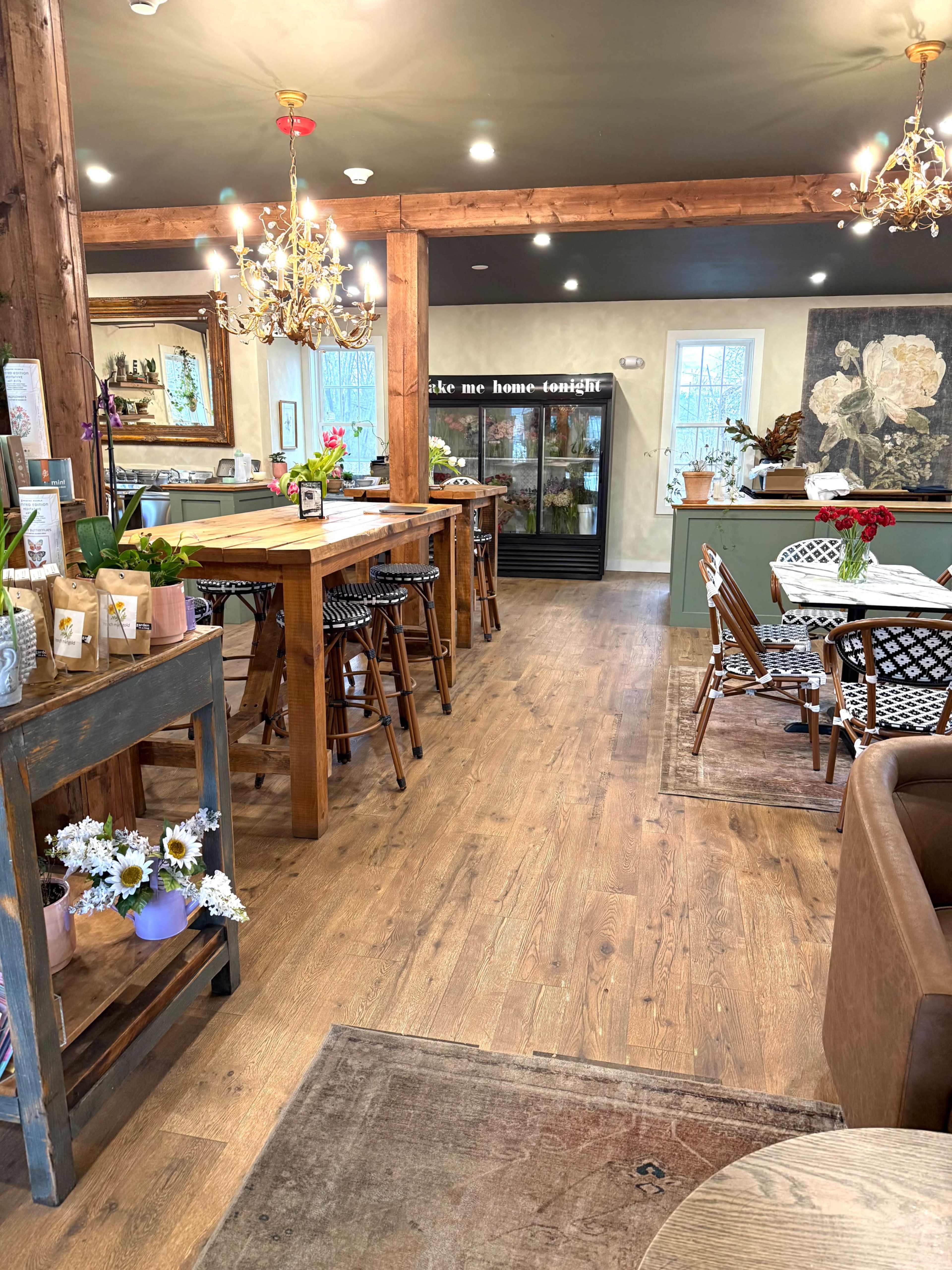 The image shows a cozy cafe with wooden tables, decorative lighting, and a display case along one wall, featuring plants and flowers.
