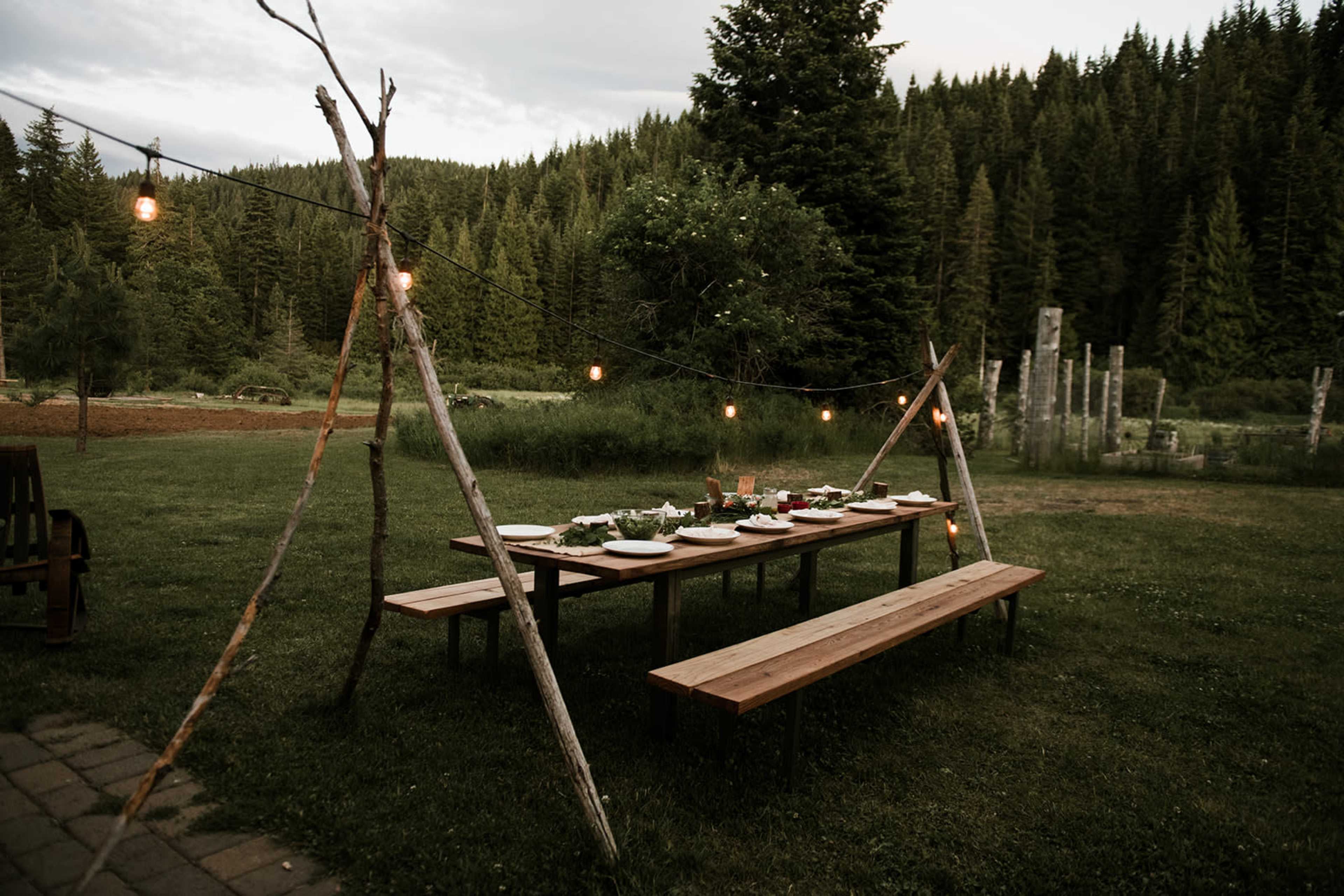A long wooden dining table set with plates and food is positioned outdoors under string lights amidst a forested landscape.