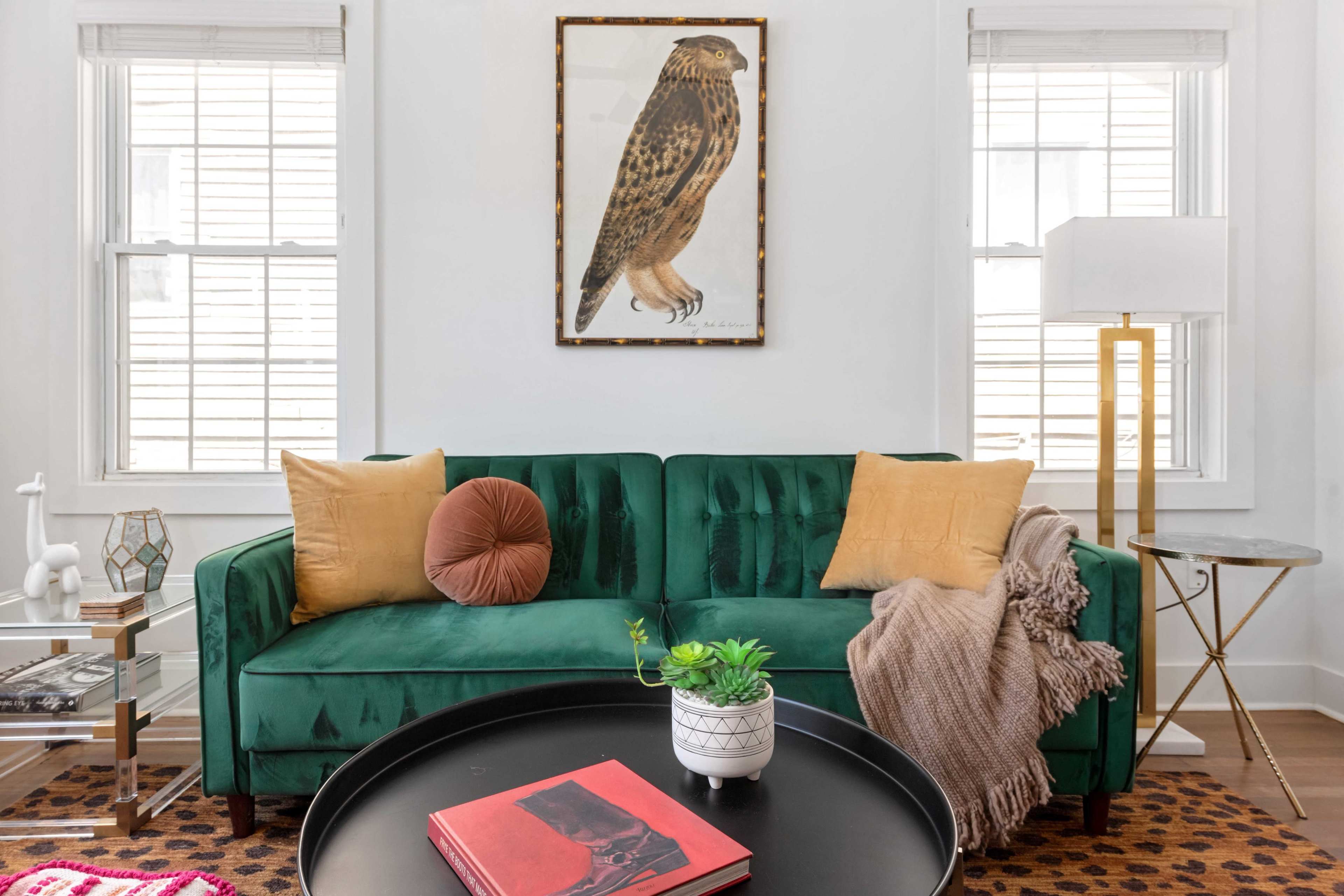 The image shows a modern living room featuring a green velvet sofa with decorative pillows, a round coffee table with books, and a large framed artwork of a bird on the wall.