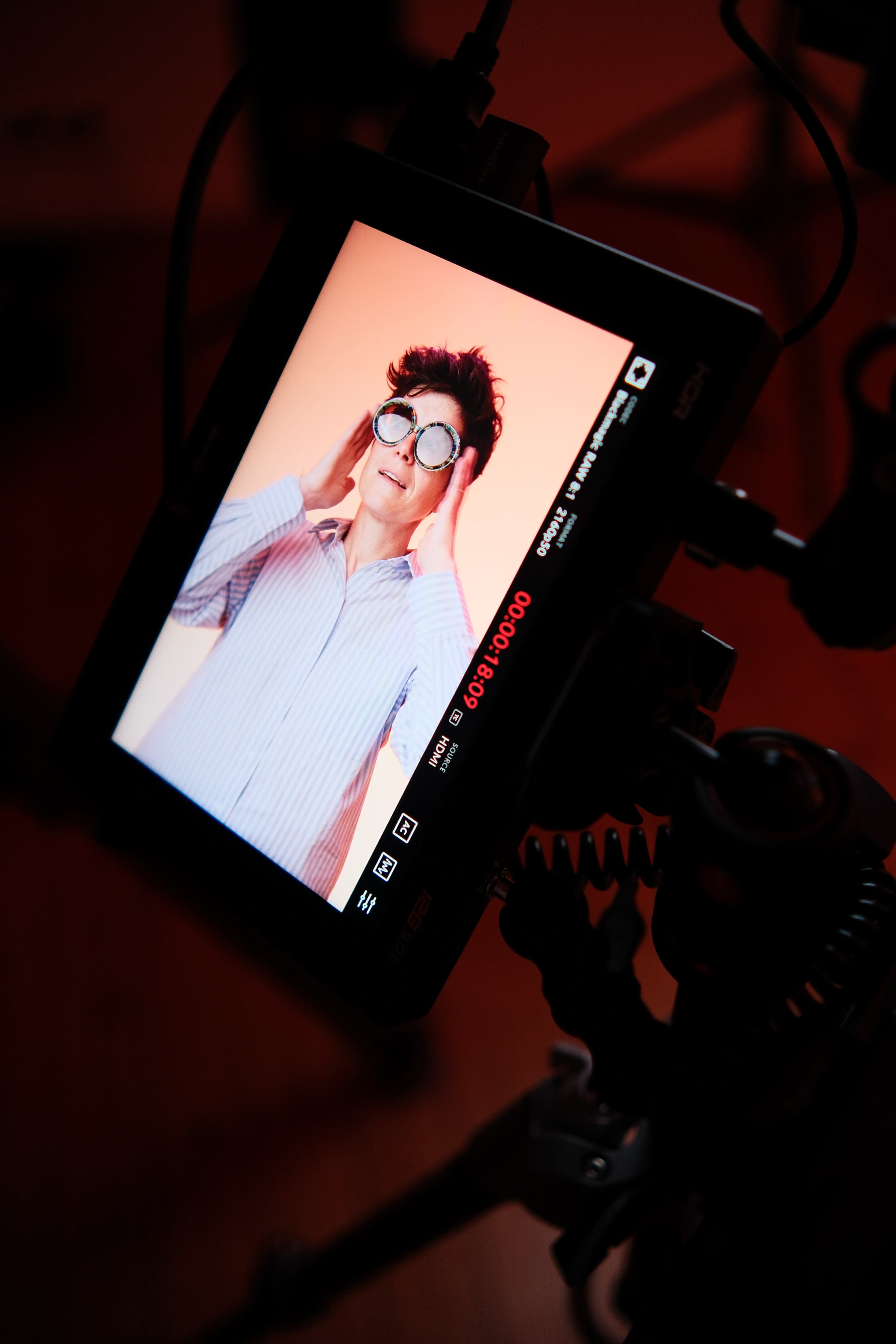A person wearing sunglasses poses while looking at a monitor displaying their image in a studio setting.