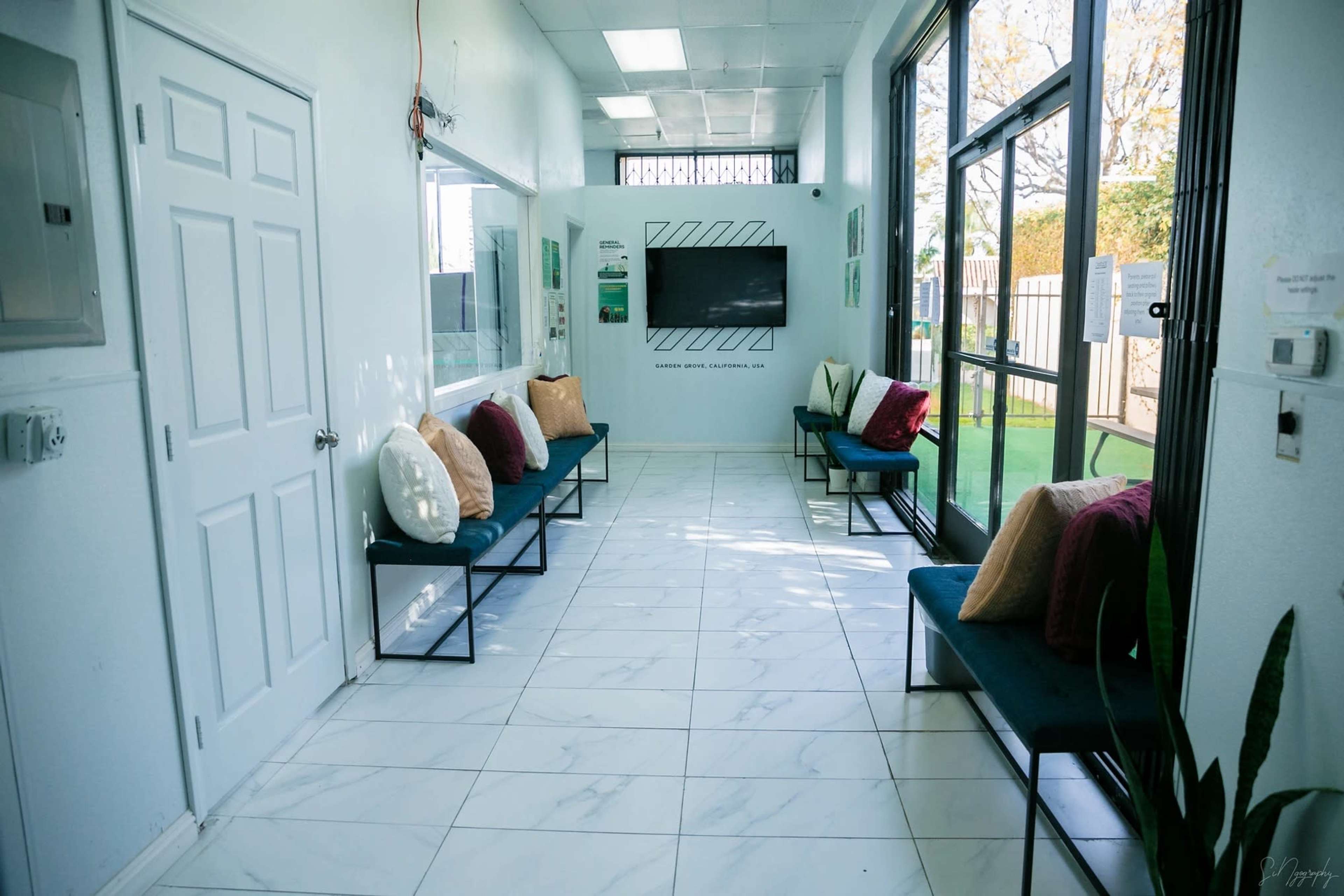 The image shows a waiting area with white walls, tiled flooring, and benches featuring colorful cushions, along with a television mounted on the wall.