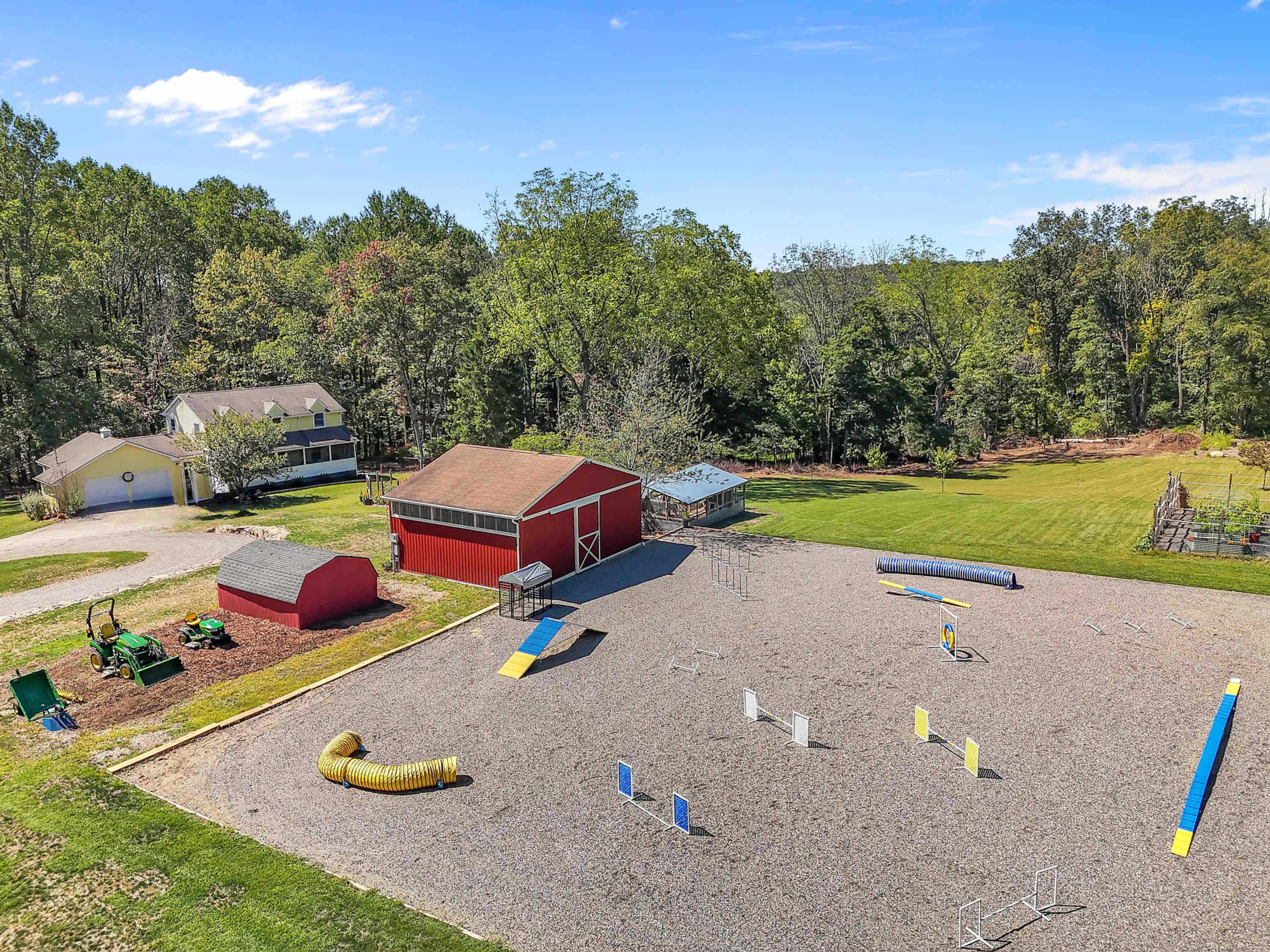 The image shows a gravel-covered area with various agility equipment for dogs, a red barn, and a large grassy space surrounded by trees.