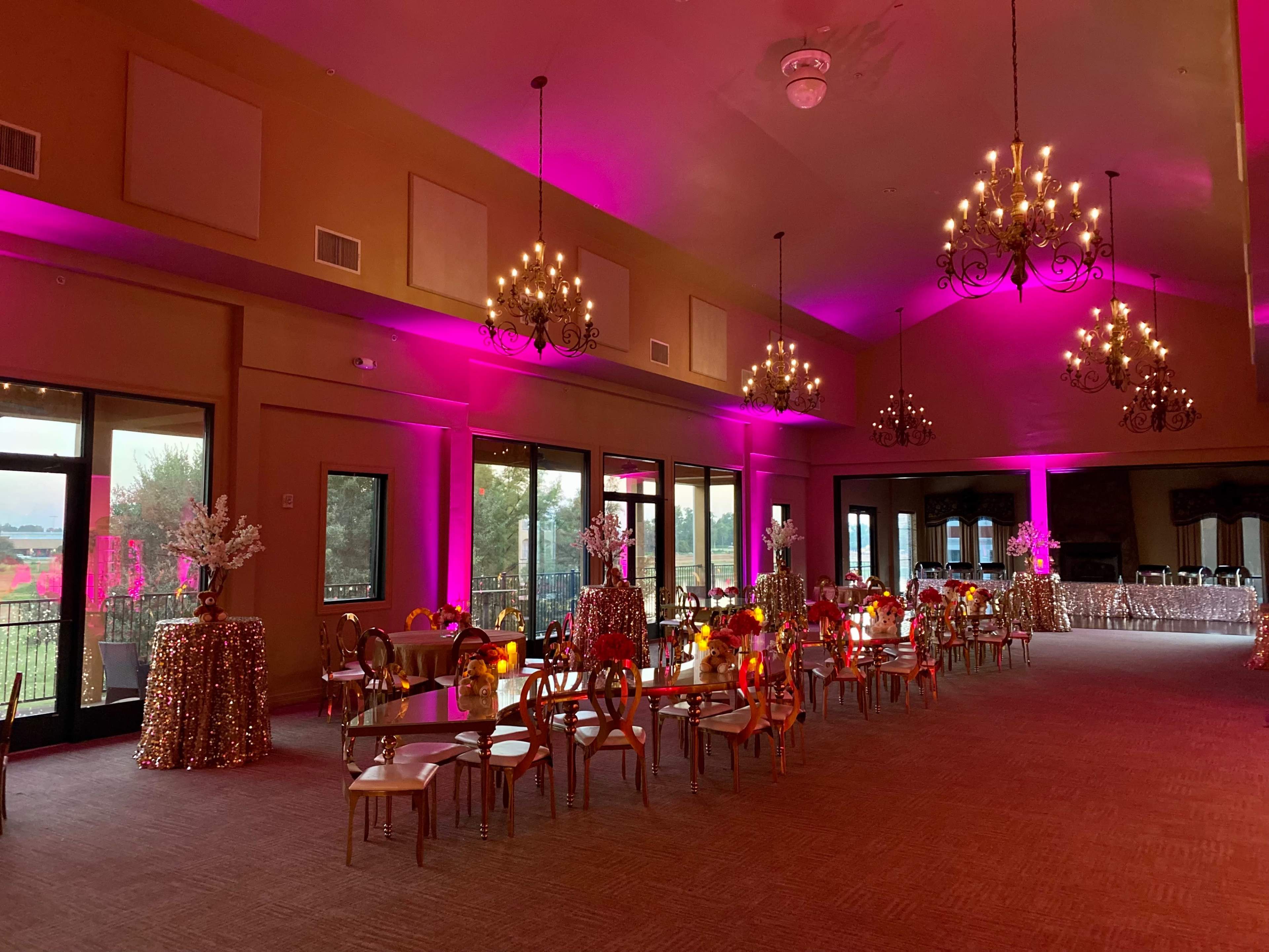 A decorated event space with pink lighting, chandeliers, and tables adorned with gold sequined tablecloths and floral arrangements.