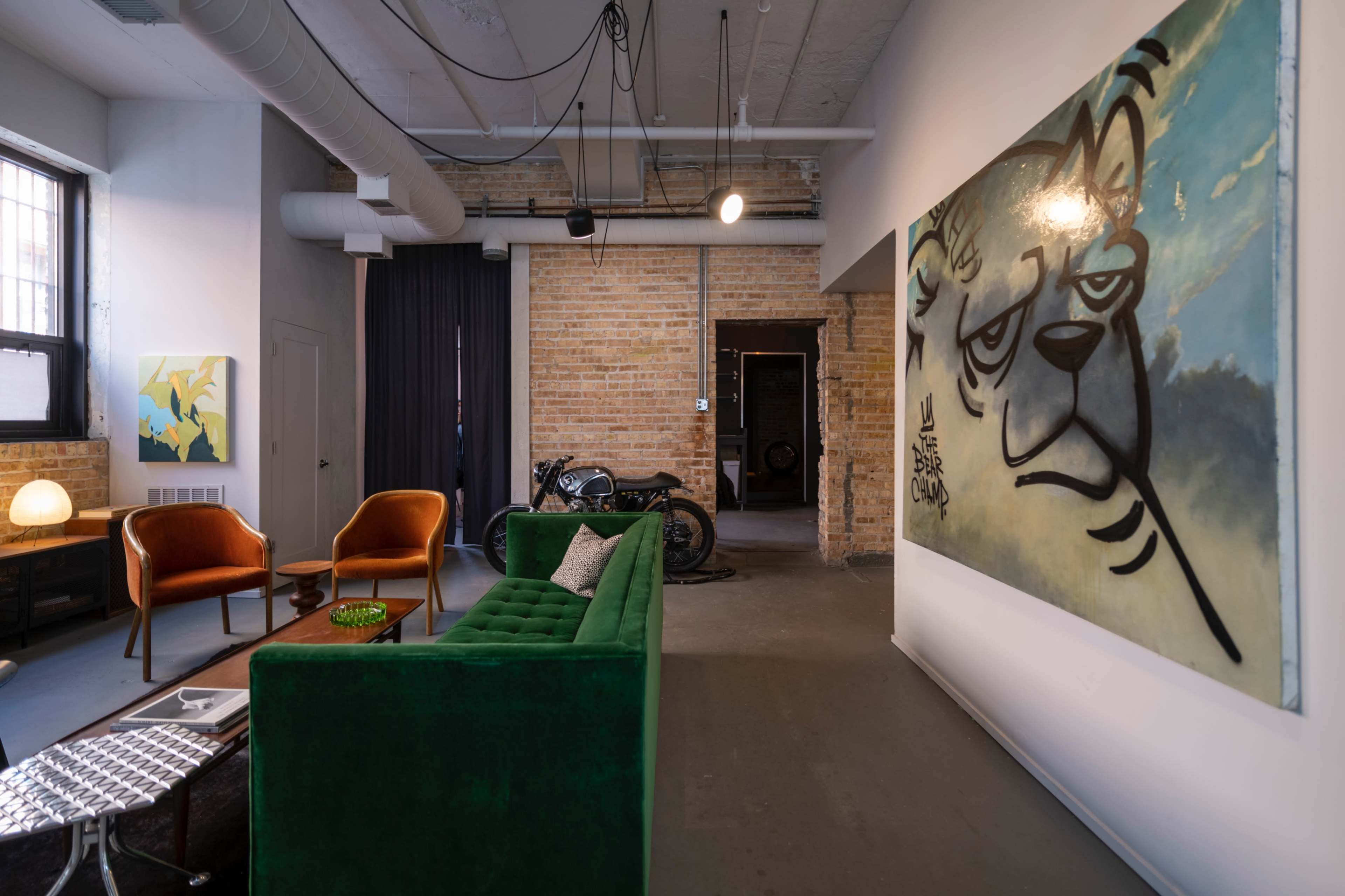 A modern, industrial-style living space with a green sofa, two orange chairs, a wall-mounted mural of a lion, and a motorcycle in the background.