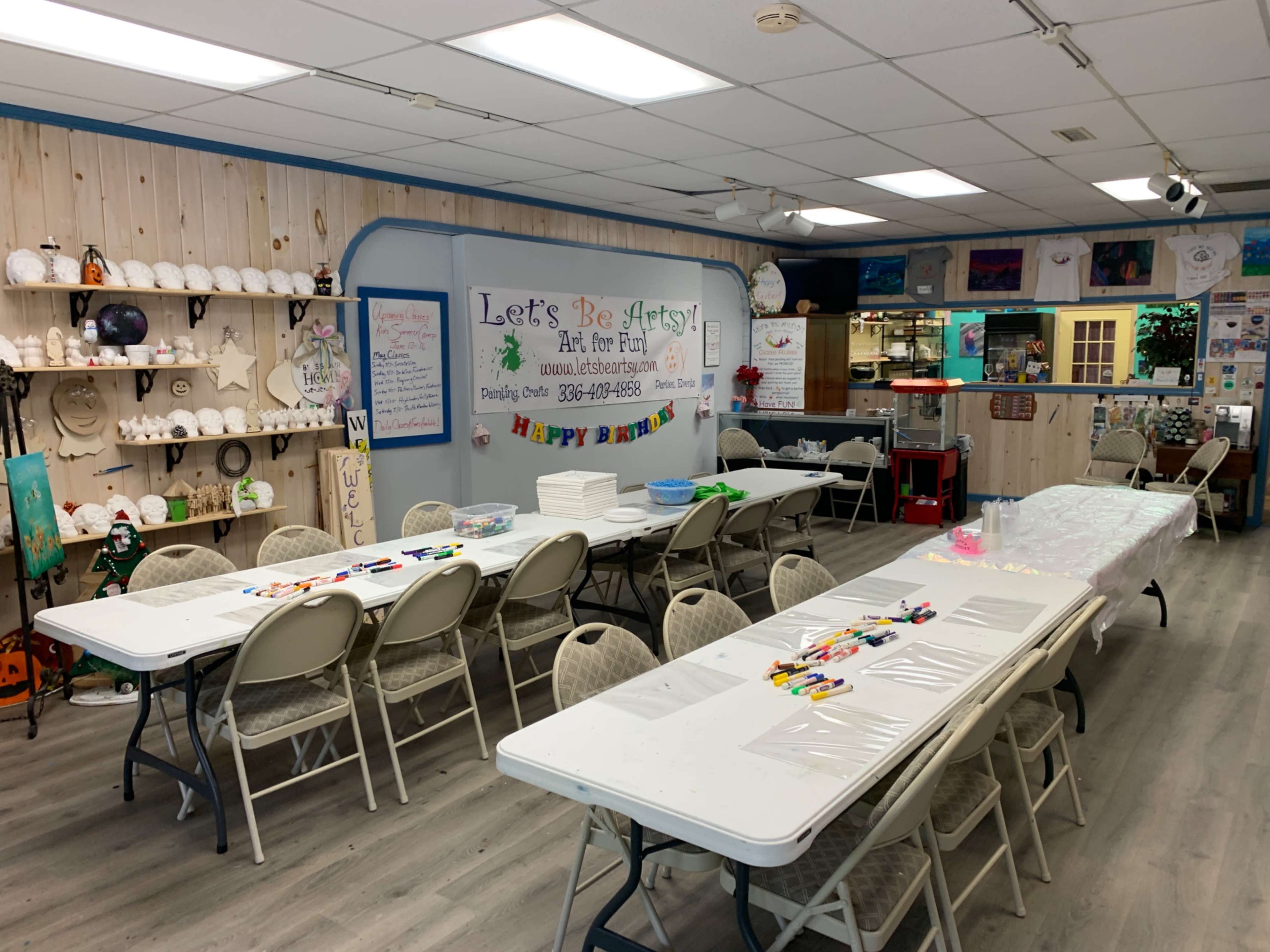A craft studio with several tables set up for painting activities, decorated with various art supplies and a colorful banner that reads "Let's Be Artsy!"