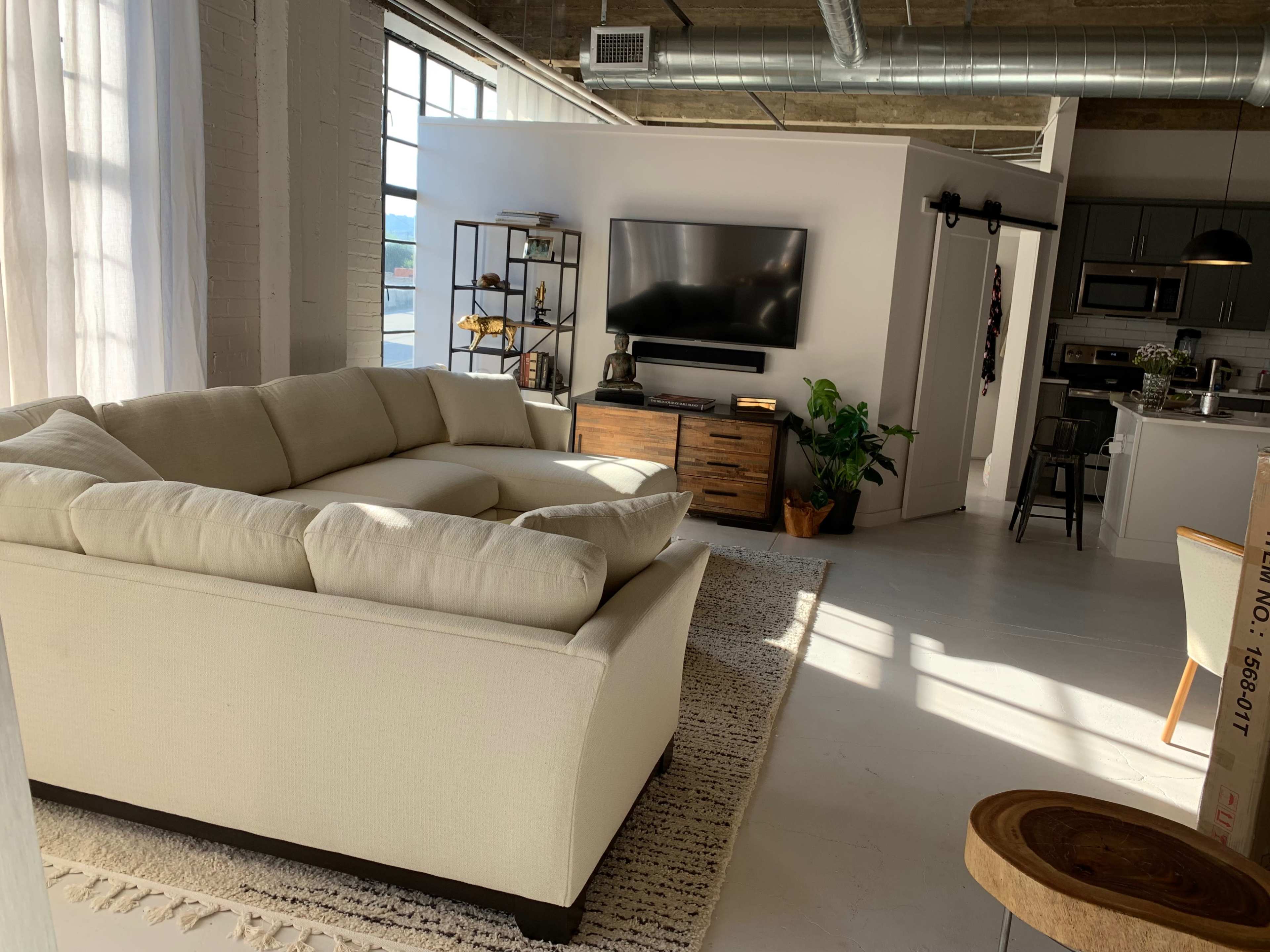 A spacious living room features a large sectional couch, a TV mounted on a wall, and an open kitchen area in the background.