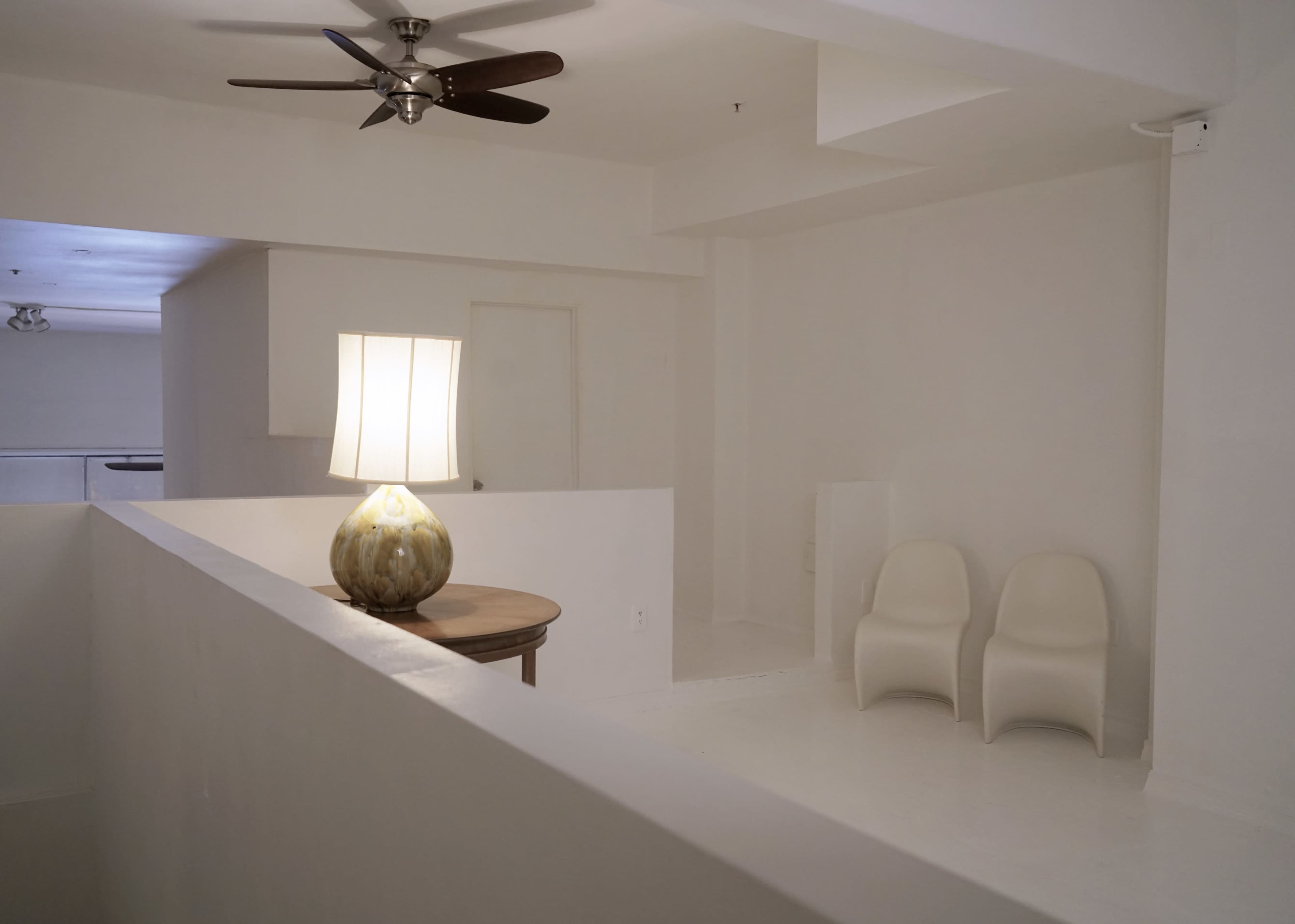 A minimalist interior features a lamp on a table, two white chairs, and a ceiling fan in a space with white walls and flooring.