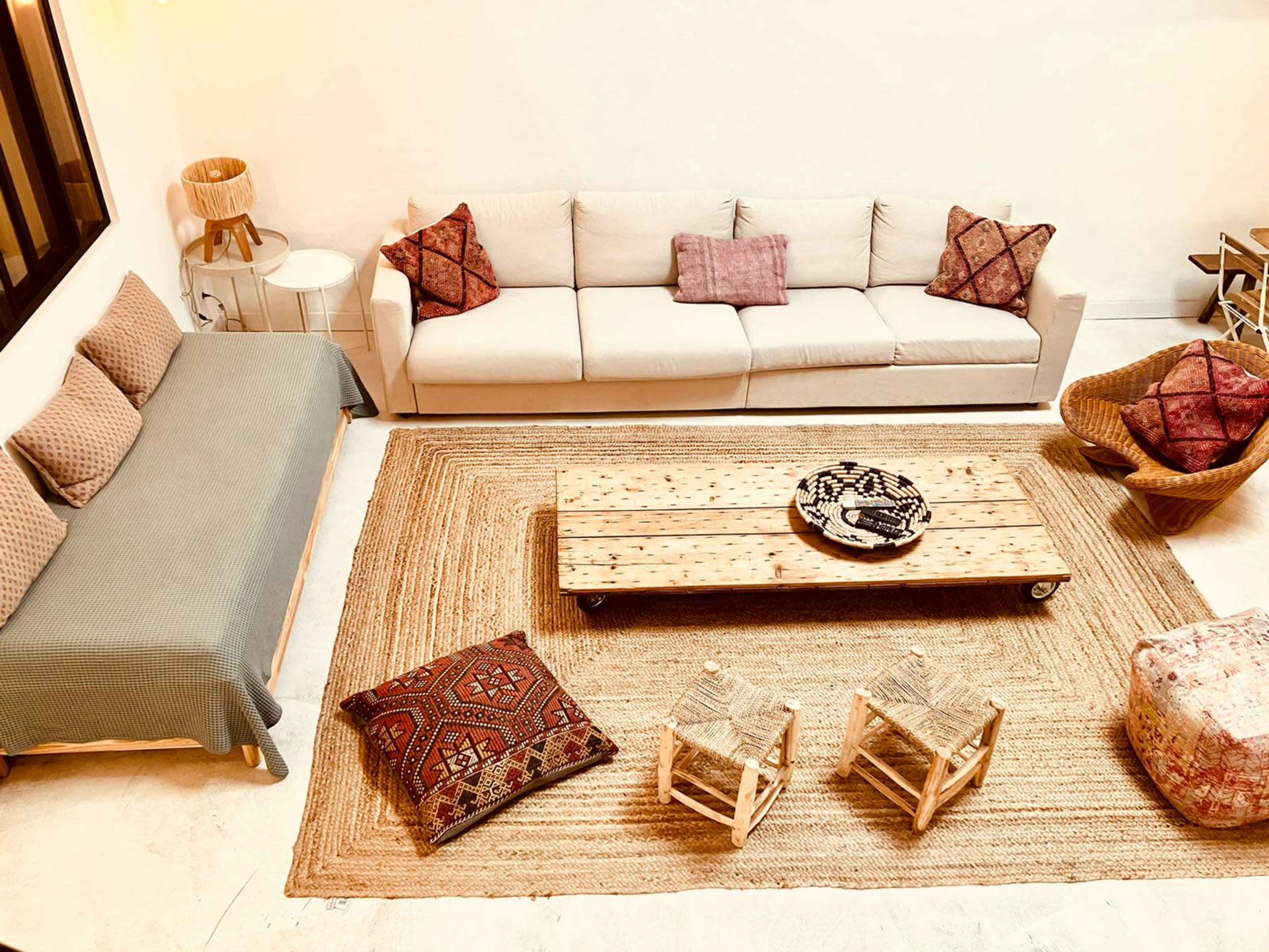 The image shows a cozy living room with a light-colored sofa, a wooden coffee table, and several seating areas adorned with patterned cushions and a woven rug.