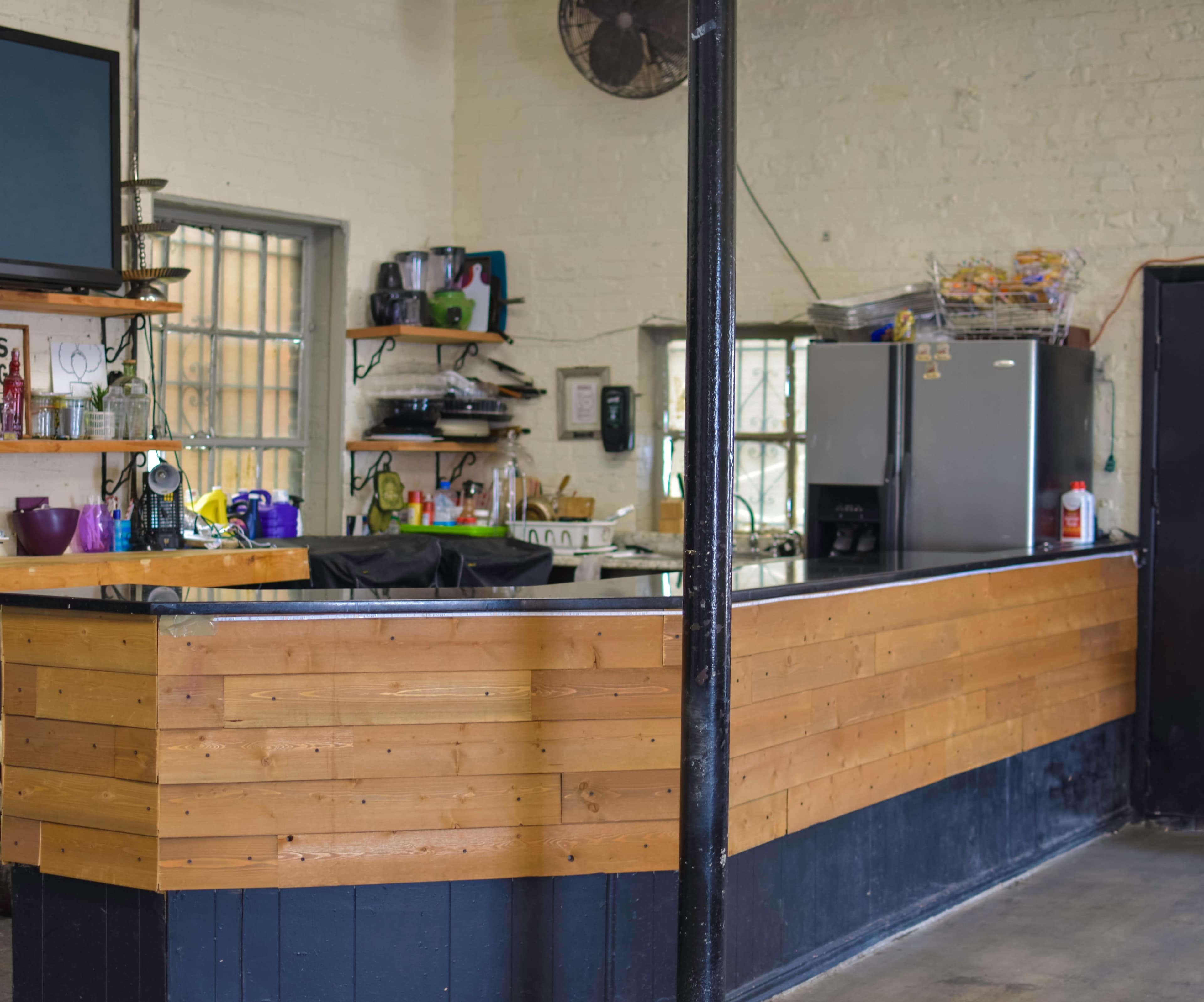 The image shows a kitchen bar area with a wooden countertop, shelves displaying various kitchen items, and a refrigerator in the background.