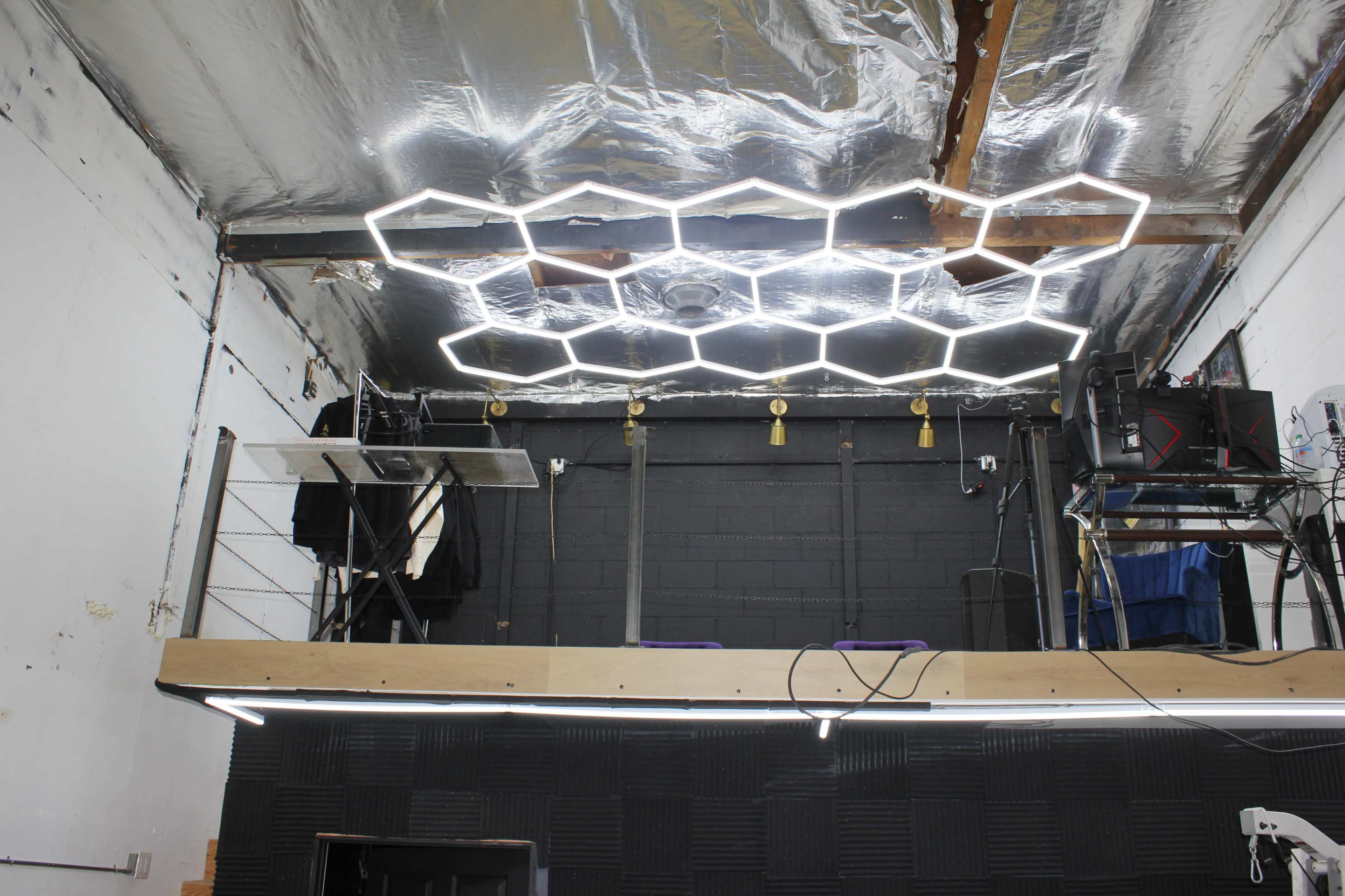 A room with a hexagonal LED light fixture on the ceiling, a raised platform with shelves, and various equipment on a black wall.