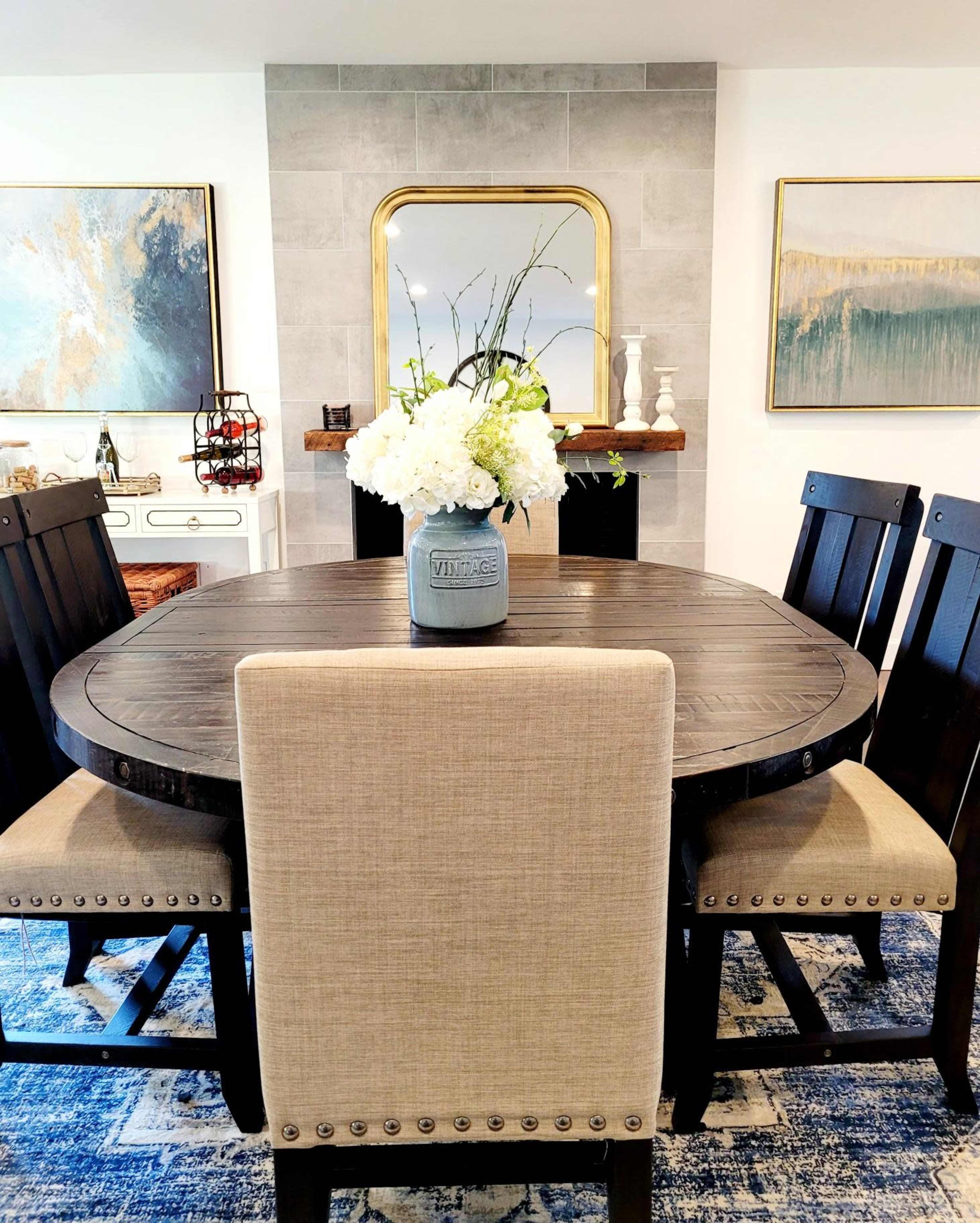 A round dining table surrounded by four upholstered chairs and decorated with a vase of flowers, set against a backdrop of a stone fireplace and framed artwork.