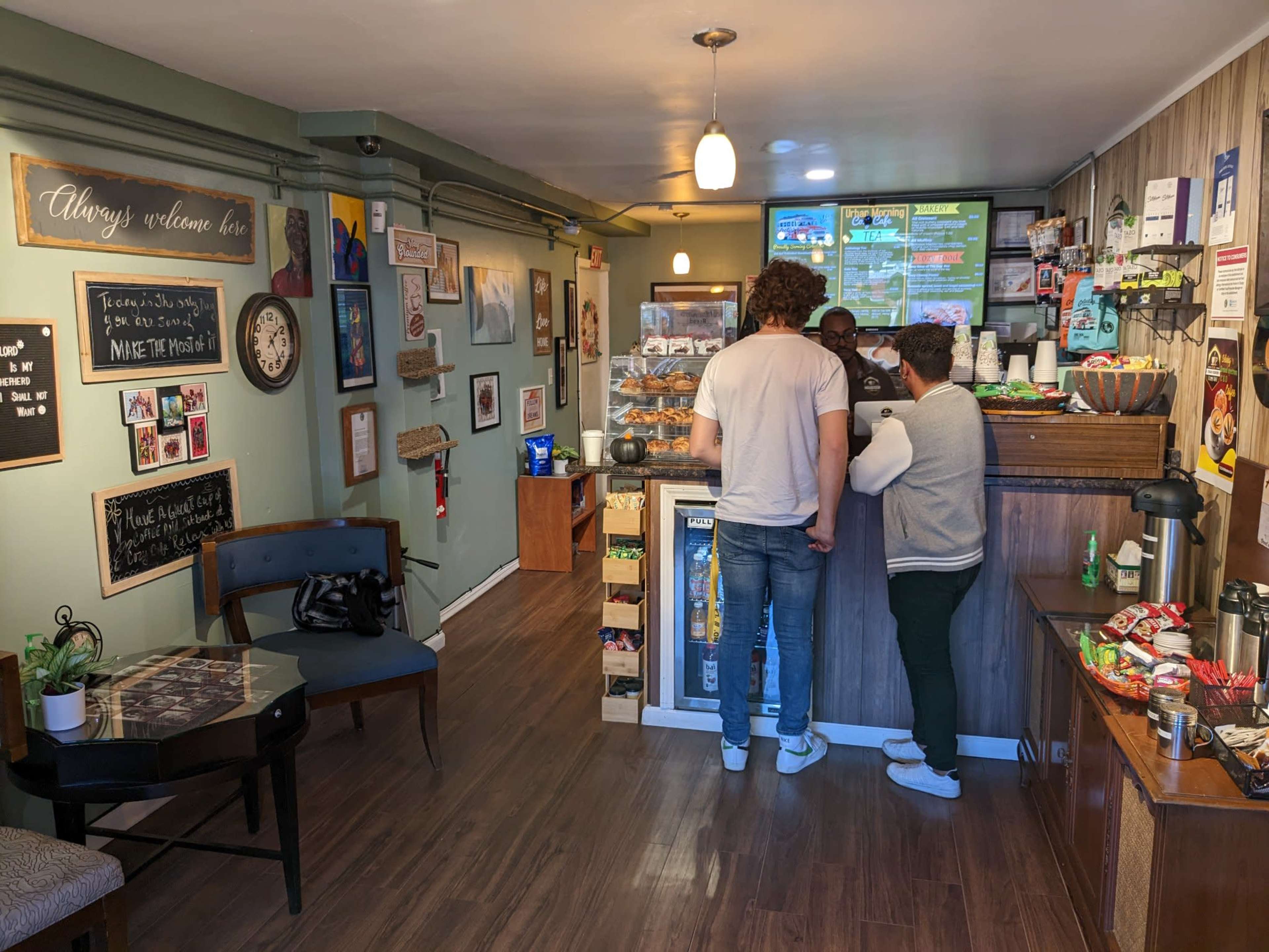 Two customers stand at the counter of a cozy cafe, engaging with the barista while a variety of snacks and beverages are displayed behind the glass case.