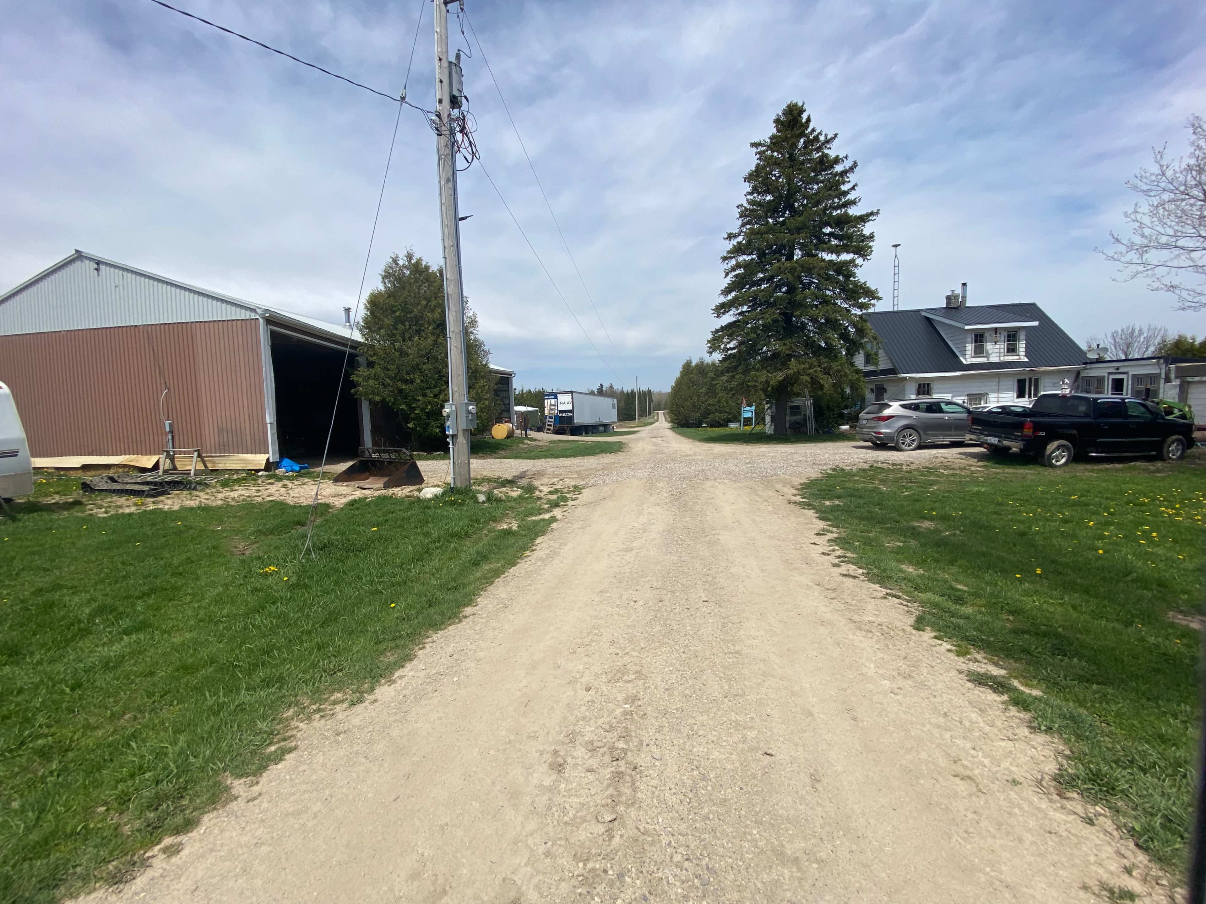A dirt road leads through a rural area with two buildings on either side, flanked by trees and parked vehicles.