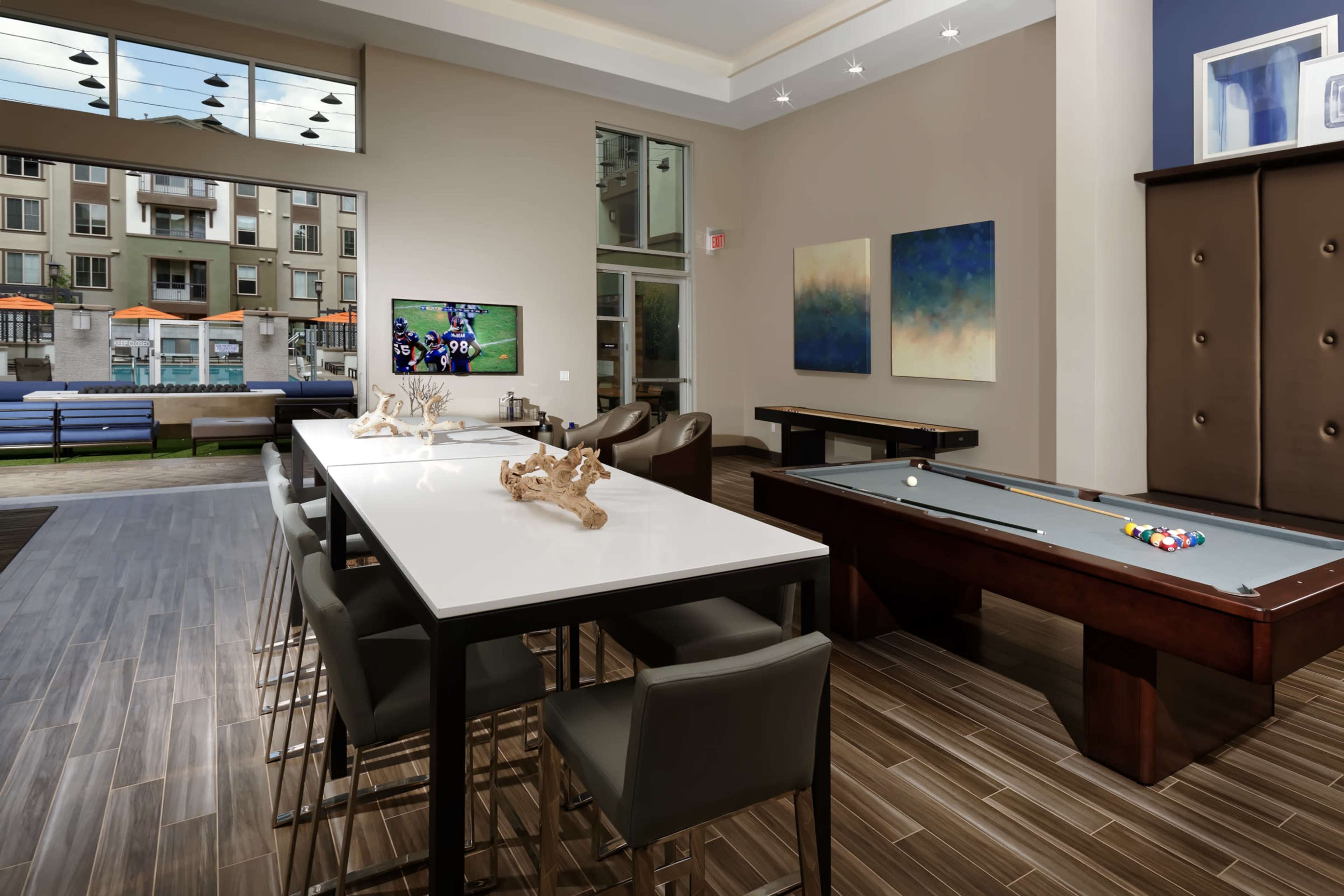 A modern lounge area featuring a pool table, a long dining table with chairs, and large windows overlooking an outdoor space with seating.