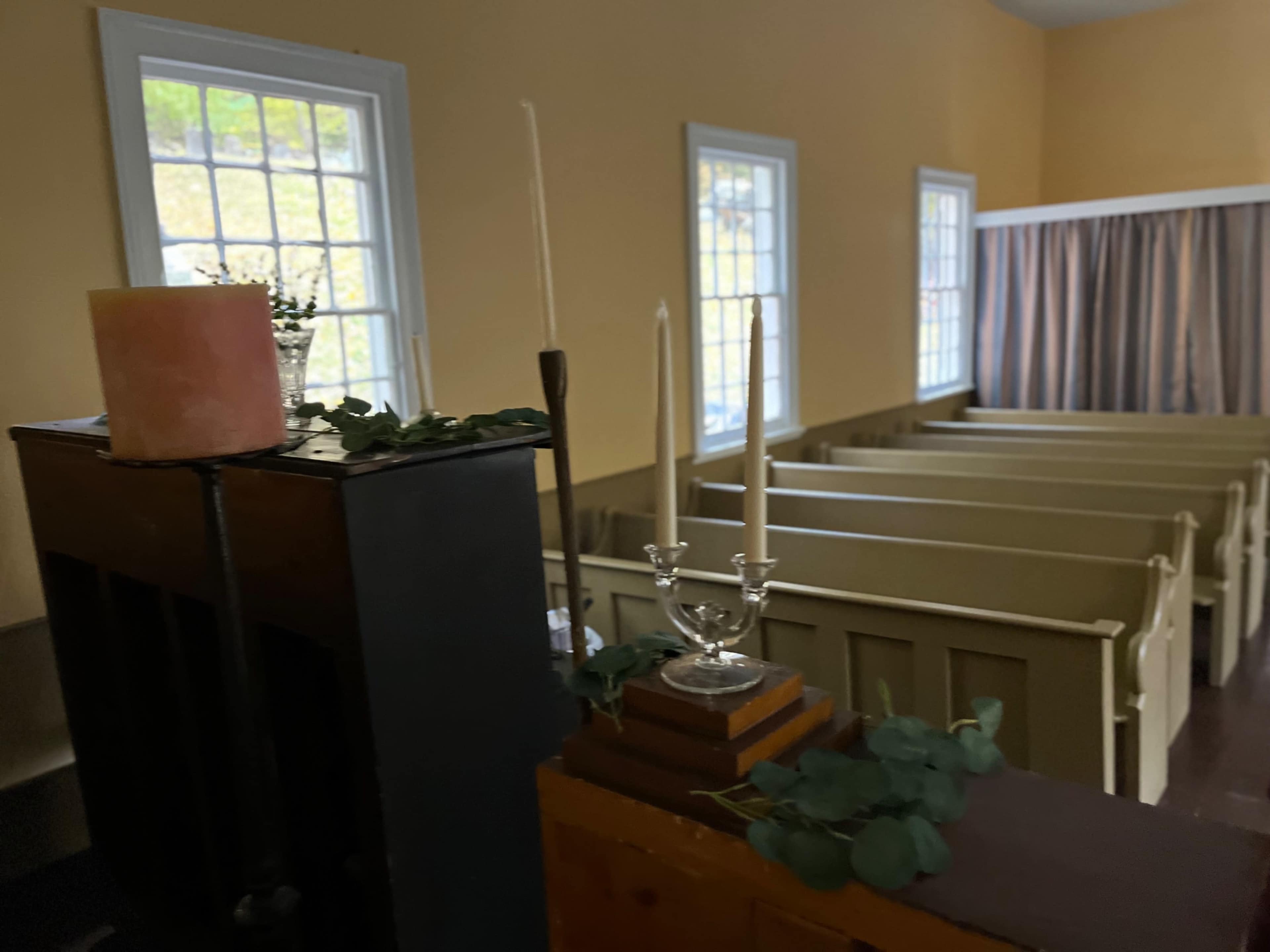 The image shows the interior of a small, empty chapel with wooden pews, a candle holder, and decorative greenery on a surface in the foreground.
