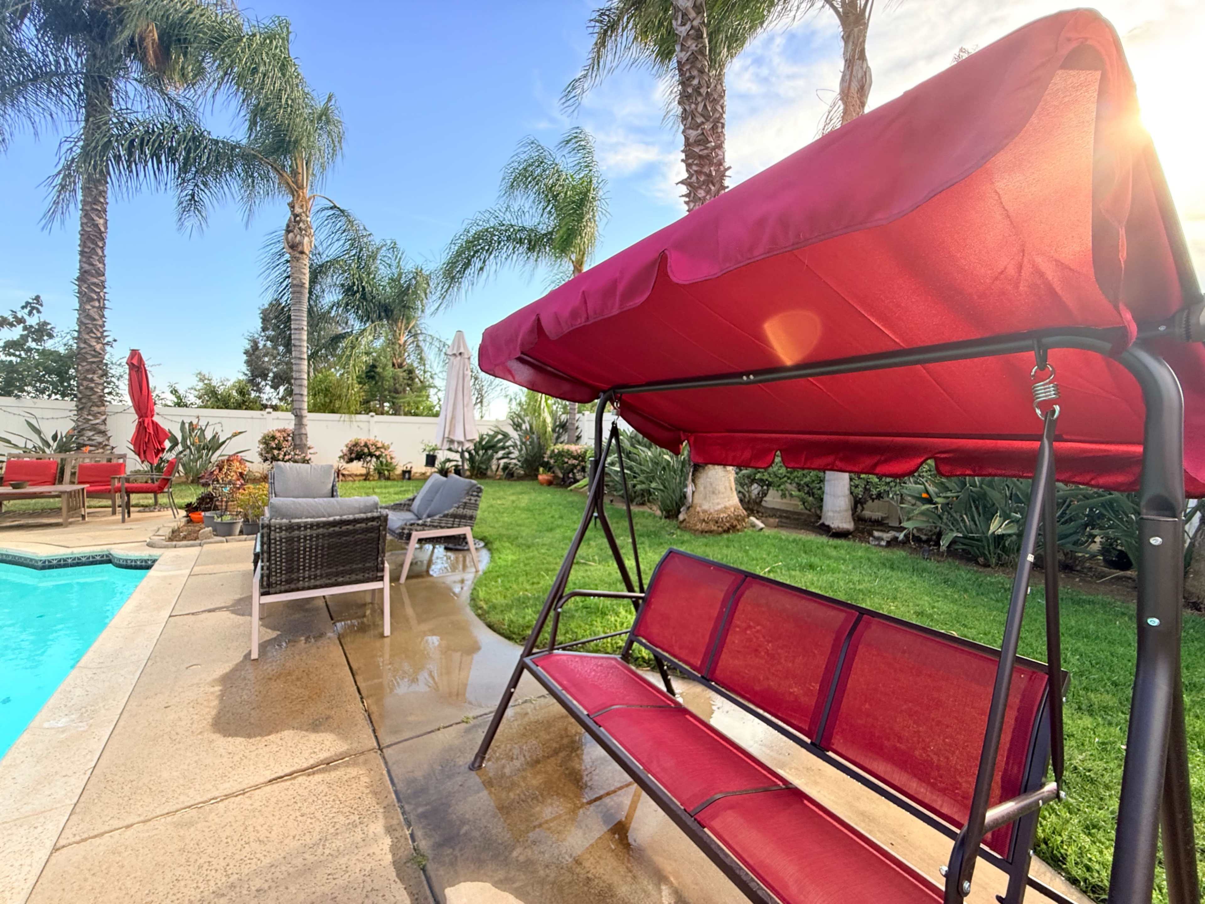 The image shows a backyard with a swimming pool, a red swing bench, and palm trees in the background.