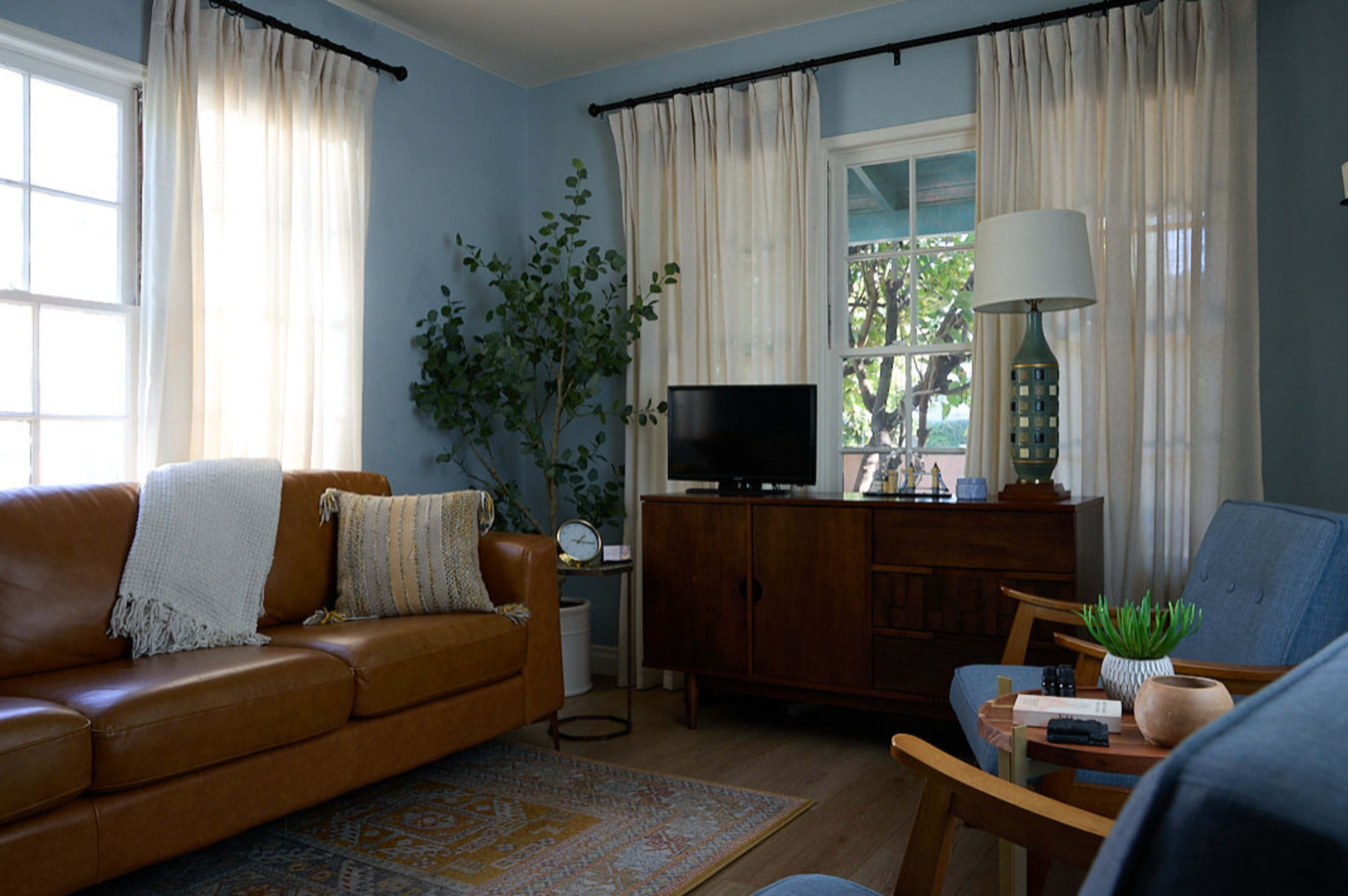 The image shows a cozy living room with a brown leather sofa, two blue chairs, a wooden cabinet, and a television near a window with light curtains.