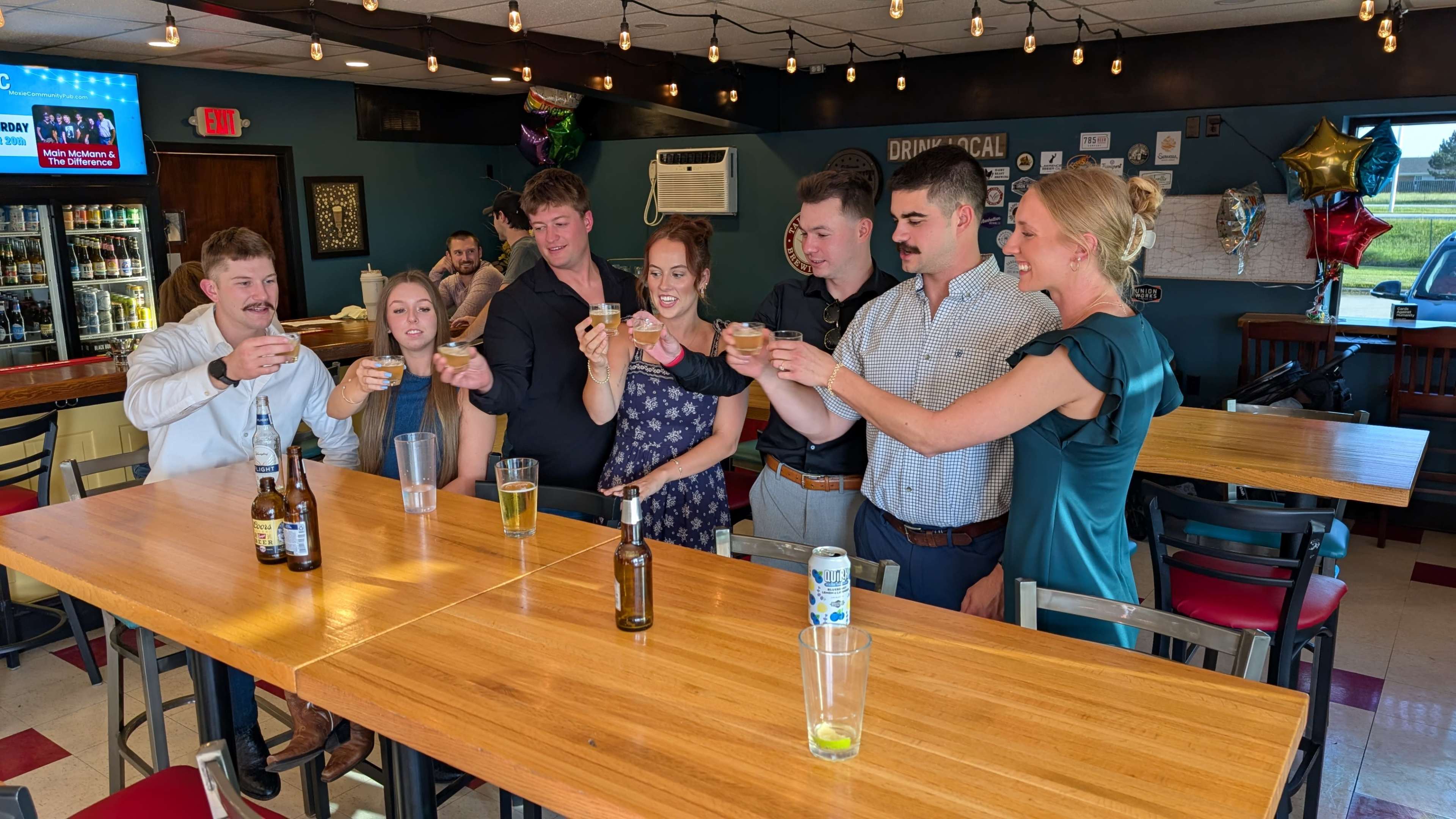A group of seven people is raising their glasses in a toast at a bar, smiling and celebrating.