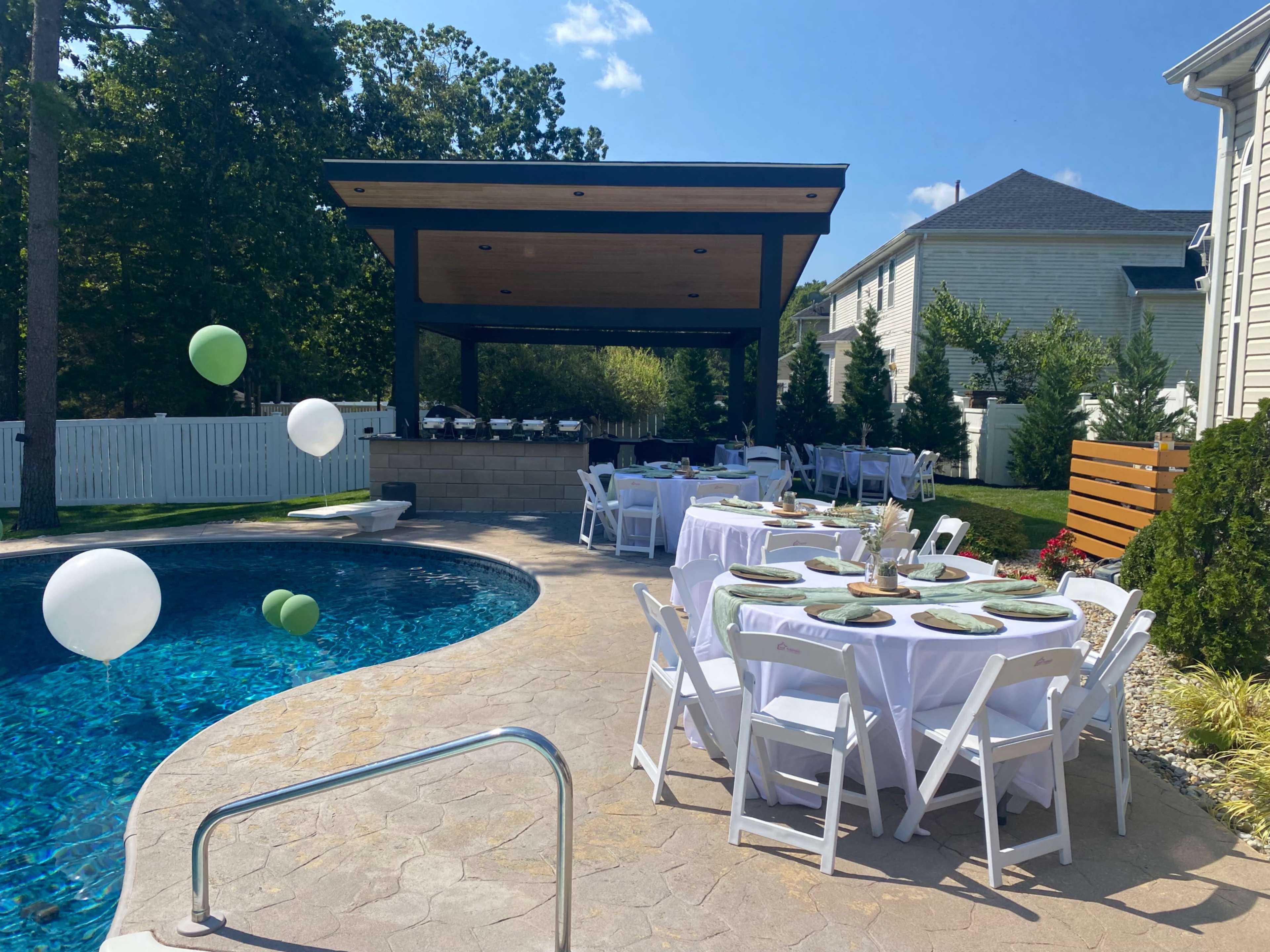 The image shows a pool area with a covered seating space and several round tables set for an outdoor event, featuring white tablecloths and balloons.