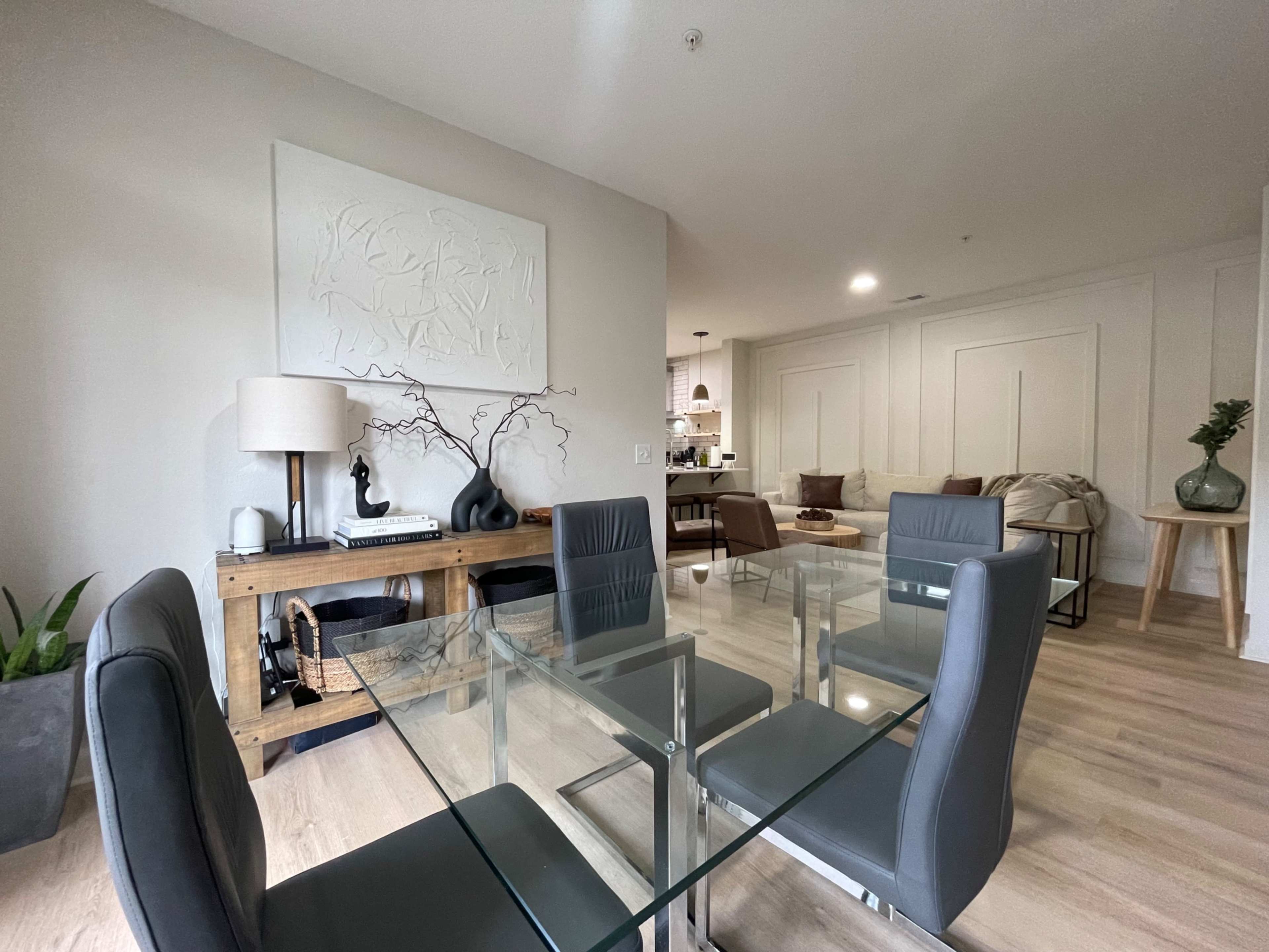 A modern dining area features a glass table with gray chairs, a wooden side table, and minimalist decor against a light-colored wall.
