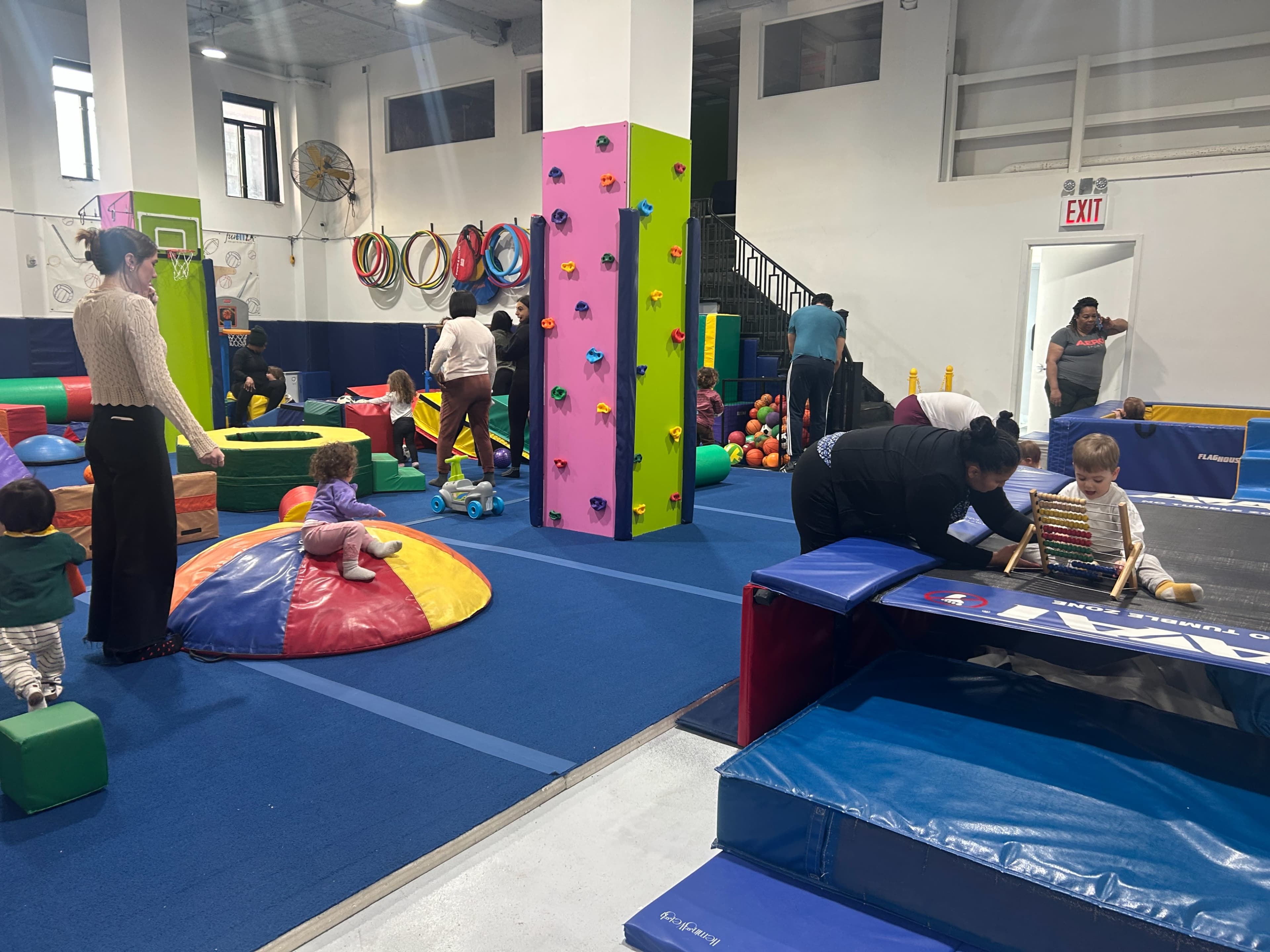 A colorful indoor play area is filled with children and adults engaging in various activities, including climbing, playing with toys, and exploring soft play structures.