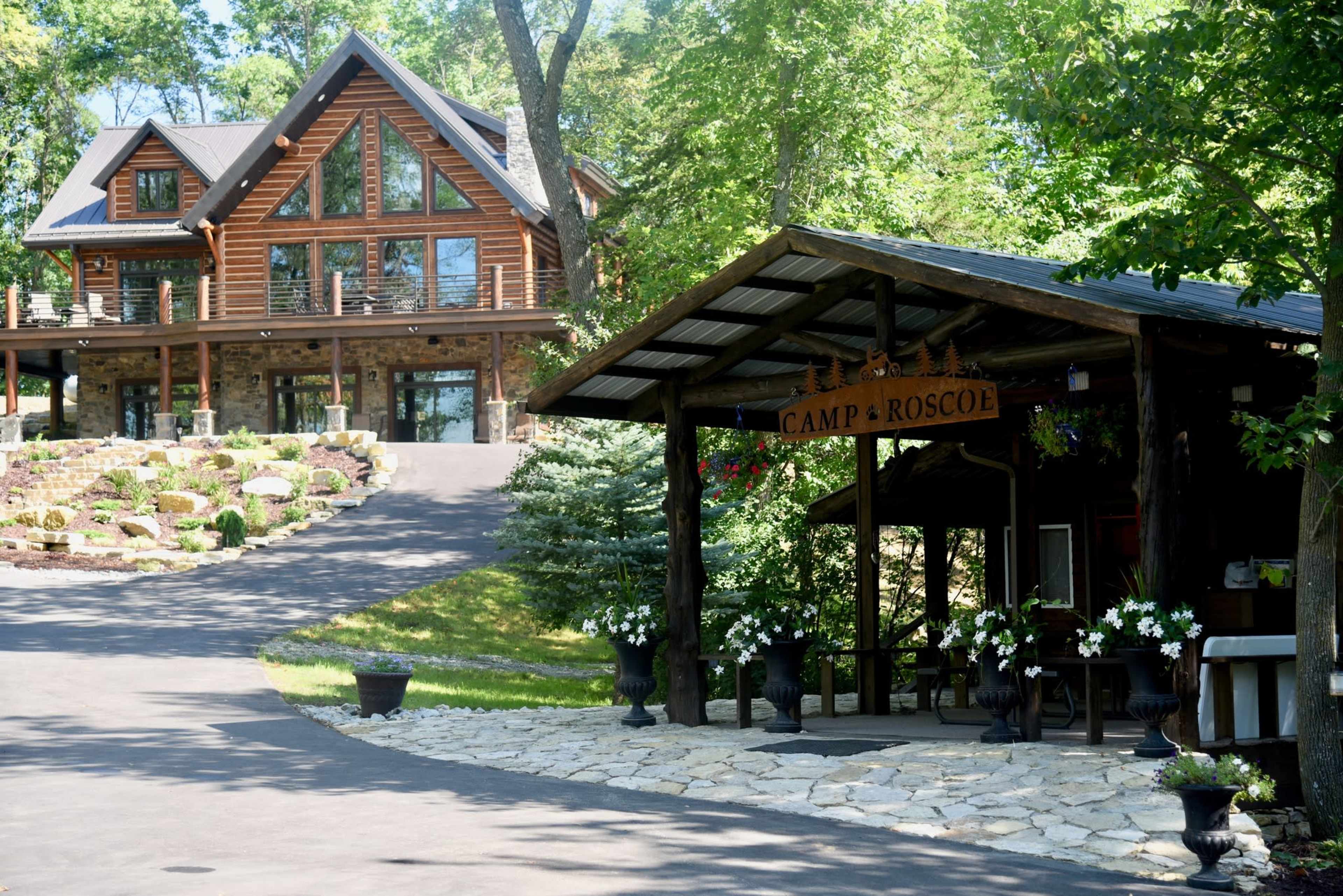 A large log cabin with multiple windows sits behind a stone pathway that leads to a rustic entrance with a sign reading "Camp Roscoe."