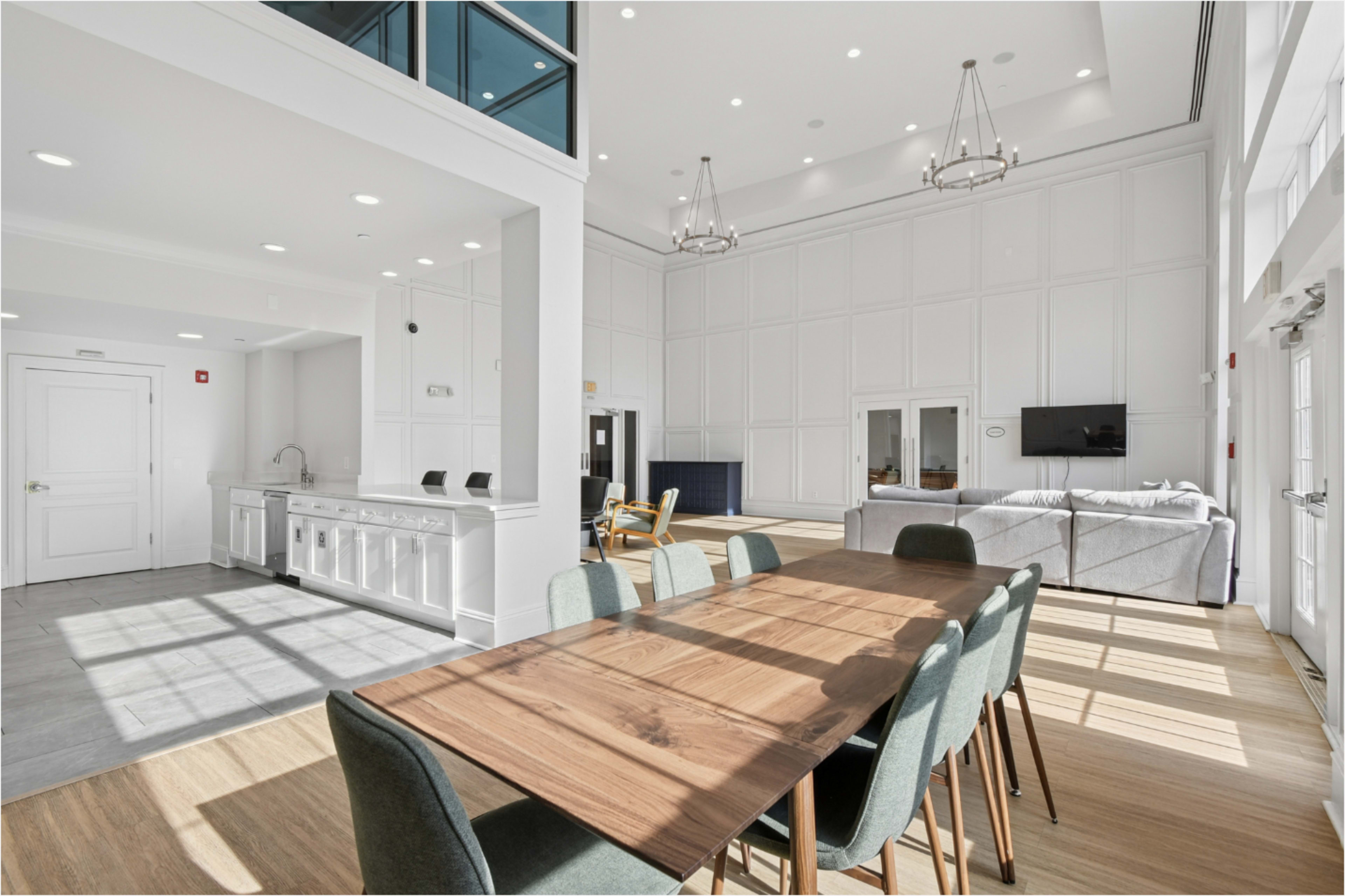 The image shows a bright, spacious common area with a large dining table, modern kitchen, and a comfortable seating arrangement.