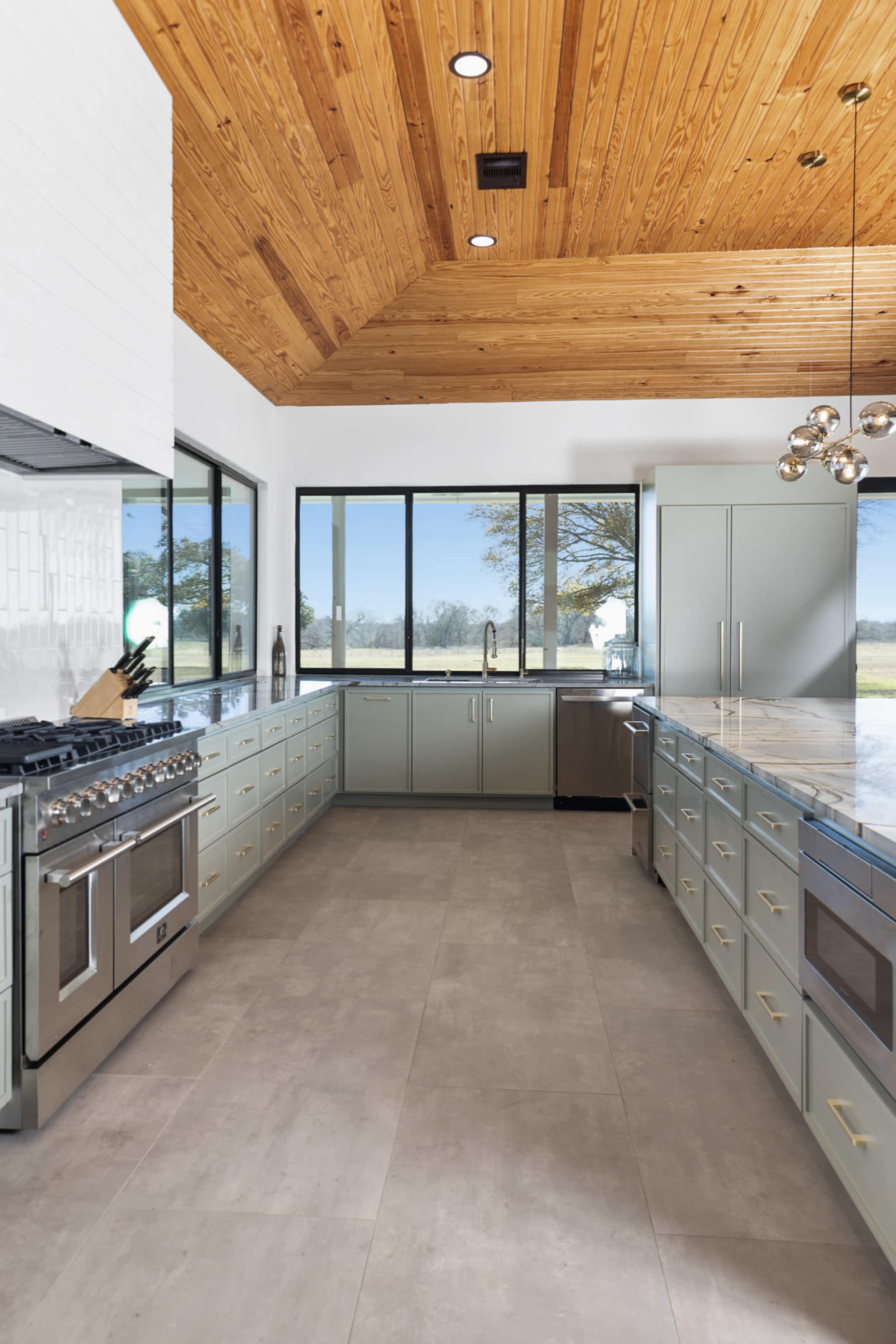 A modern kitchen features a spacious layout with light green cabinets, a marble countertop, and large windows that provide natural light.