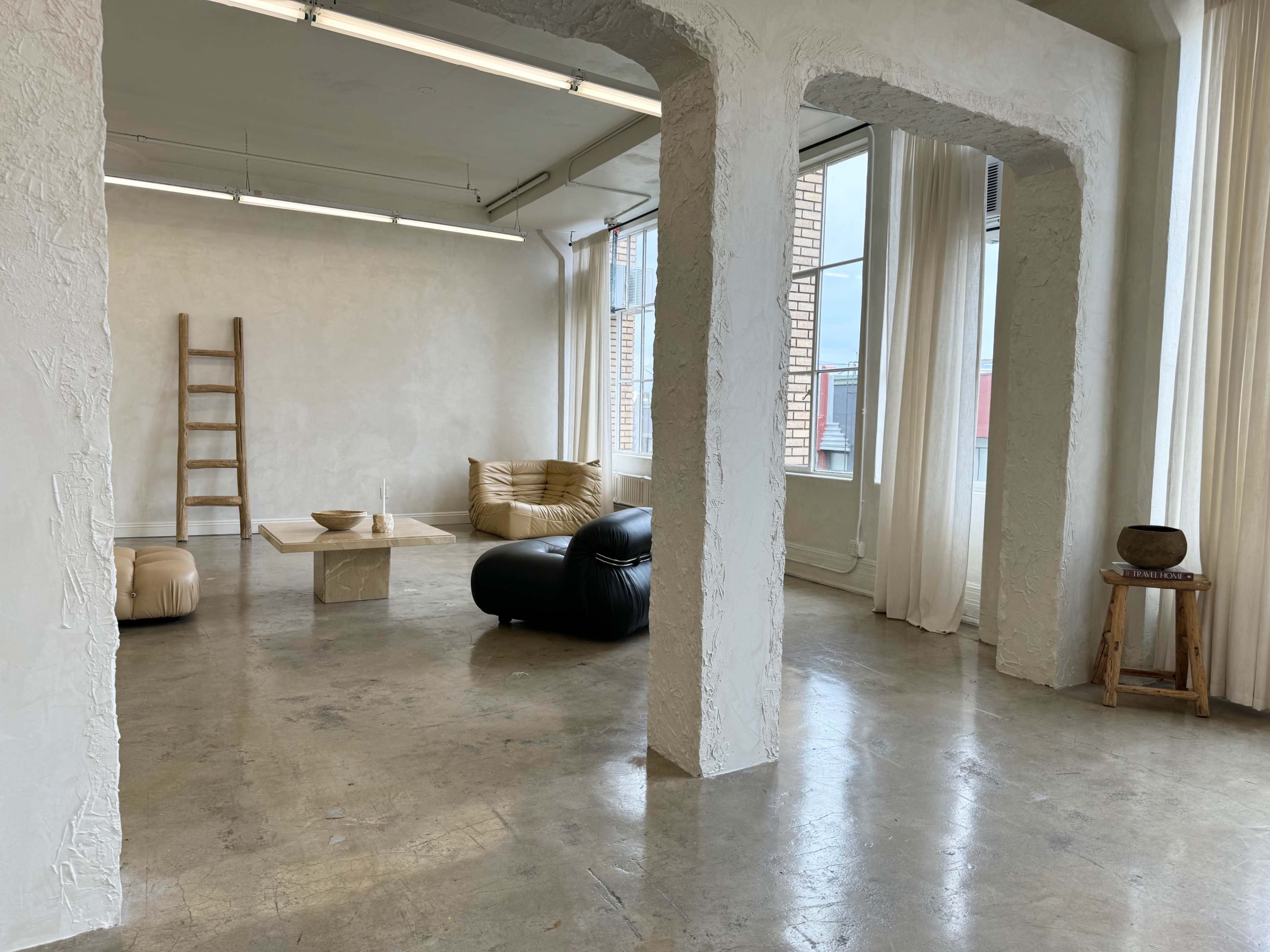 The image shows a minimalistic room with concrete floors, large windows, and a mixture of furniture including a ladder, a low table, and seating options in neutral colors.