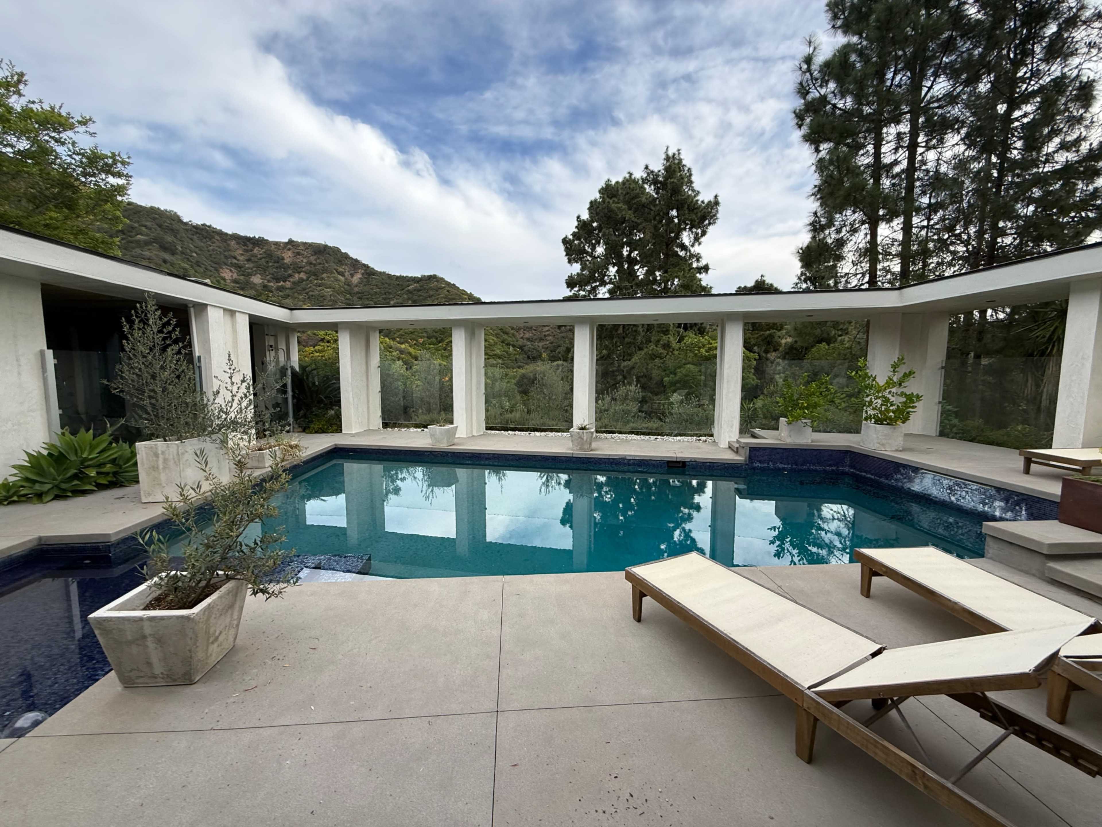 The image shows a modern outdoor swimming pool surrounded by a minimalist patio and lush greenery, with mountains in the background.