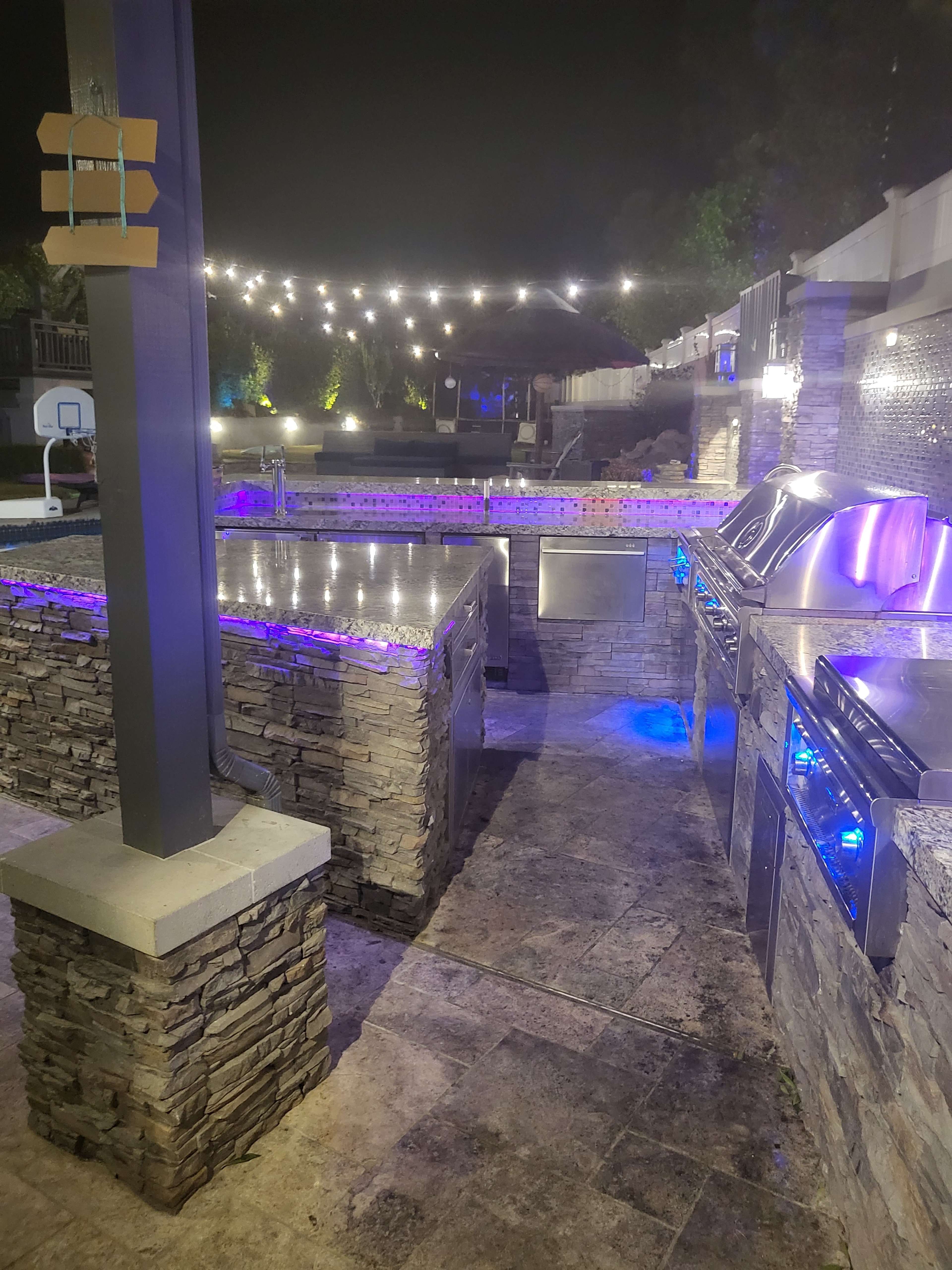 An outdoor kitchen area with stone walls, stainless steel appliances, and illuminated with purple and white lights at night.