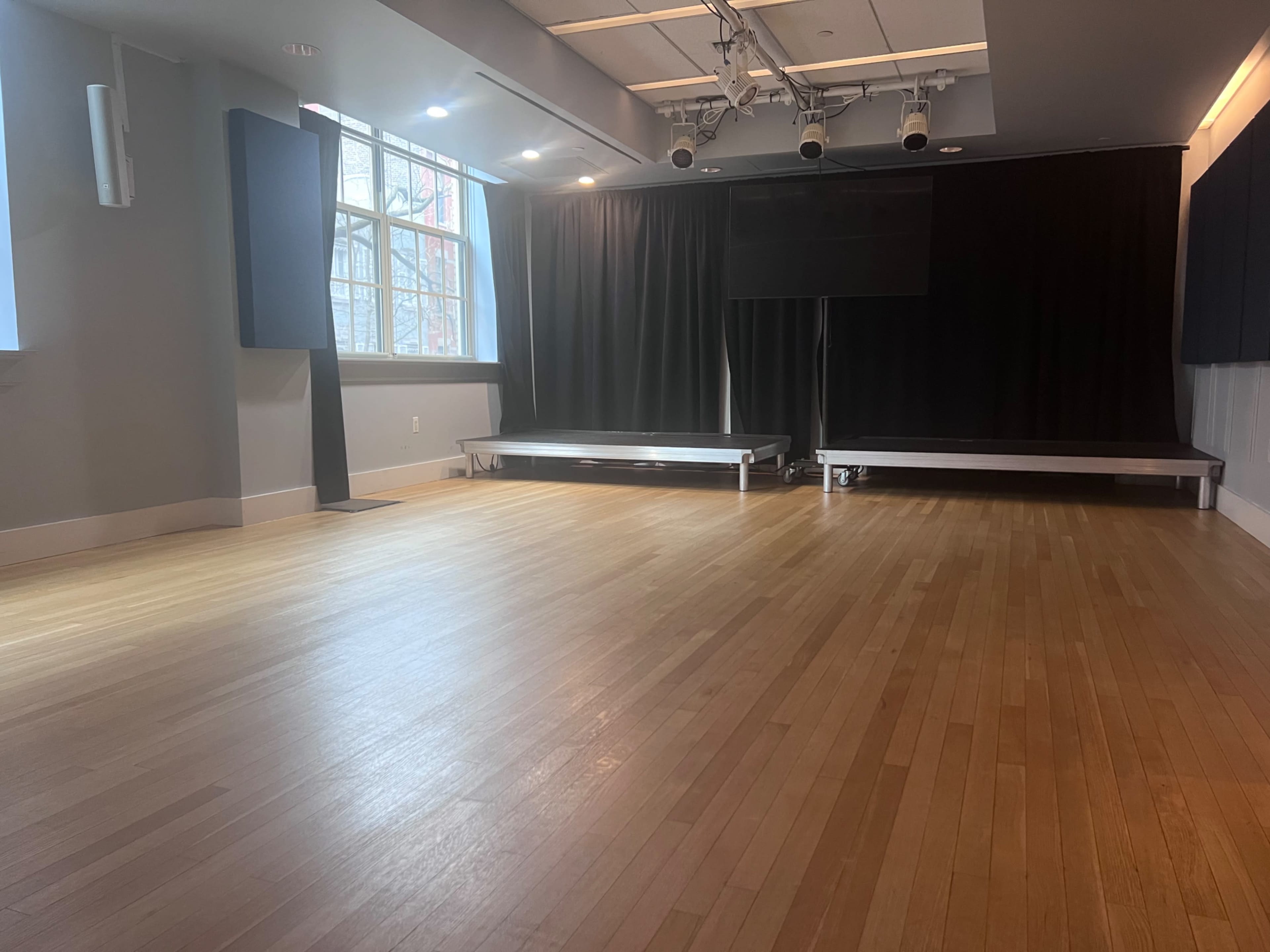 The image shows an empty studio space with wooden flooring, large windows, and dark curtains on the sides.
