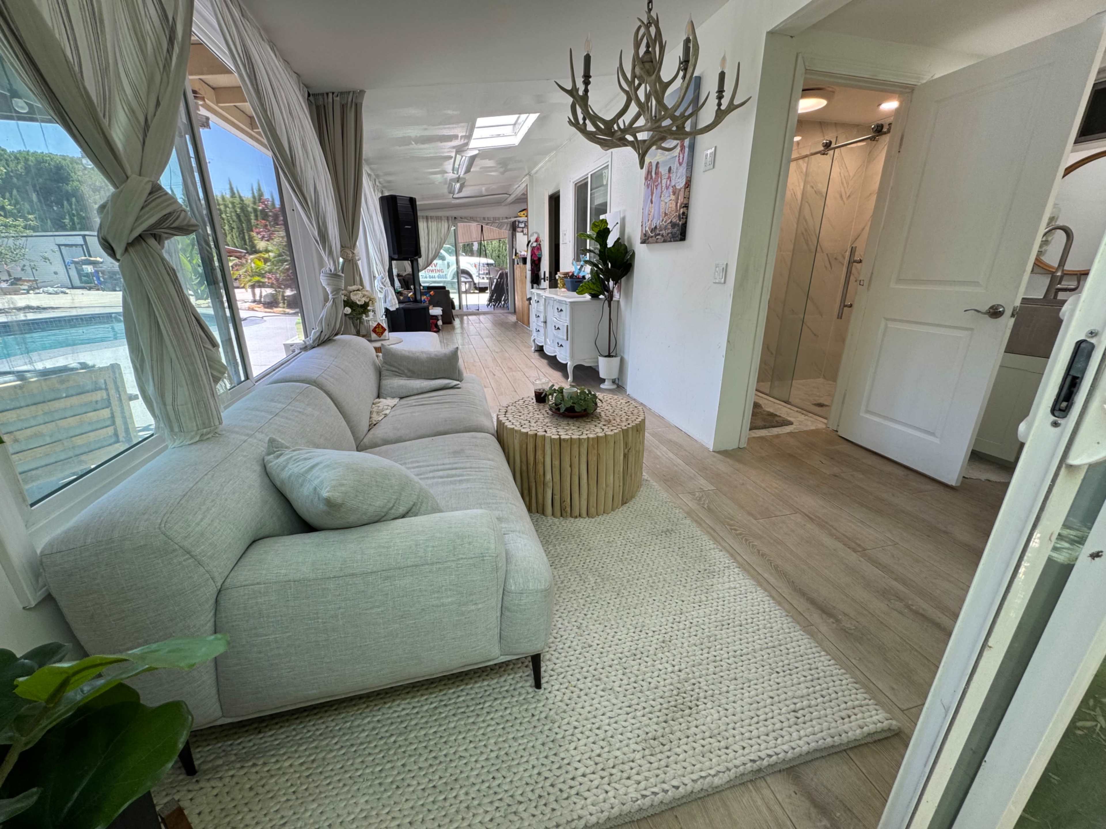 A modern living space featuring a light-colored sectional sofa, a round wooden coffee table, and a doorway leading to a bathroom, with large windows revealing a pool area outside.